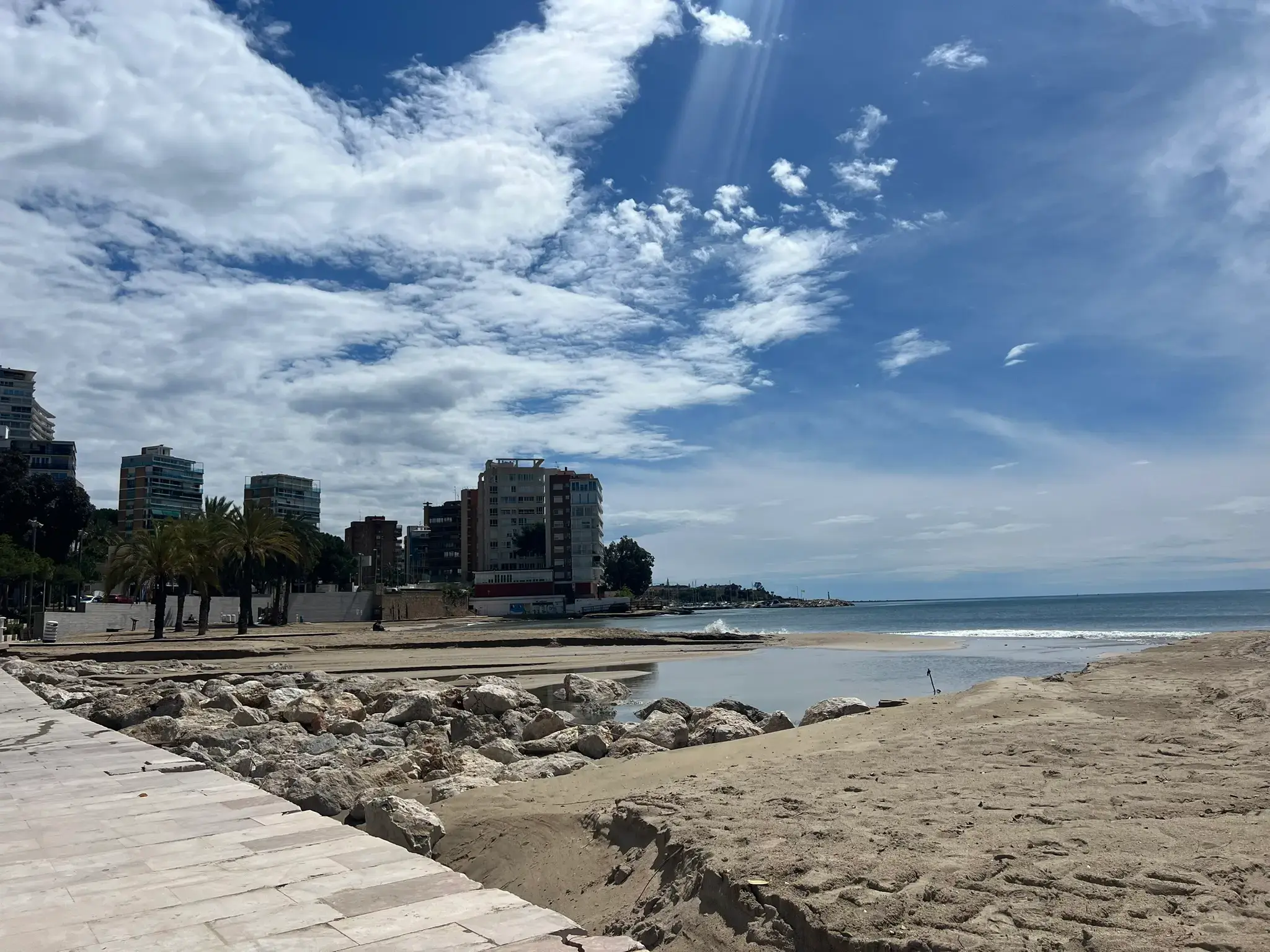 Rocky shoreline and sandy beach at Playa de la Albufereta with residential buildings and blue sky in Alicante, Spain.