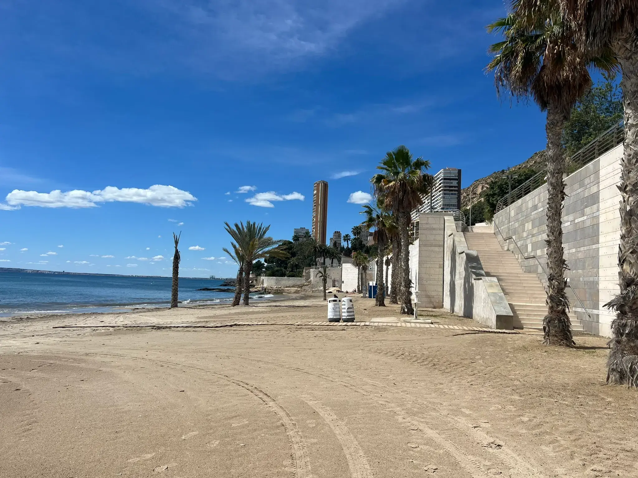 Albufereta Beach in Alicante — wide sandy shore with modern stone stairs for easy access, wooden walkways, palm trees, and the Serra Grossa hill in the background.
