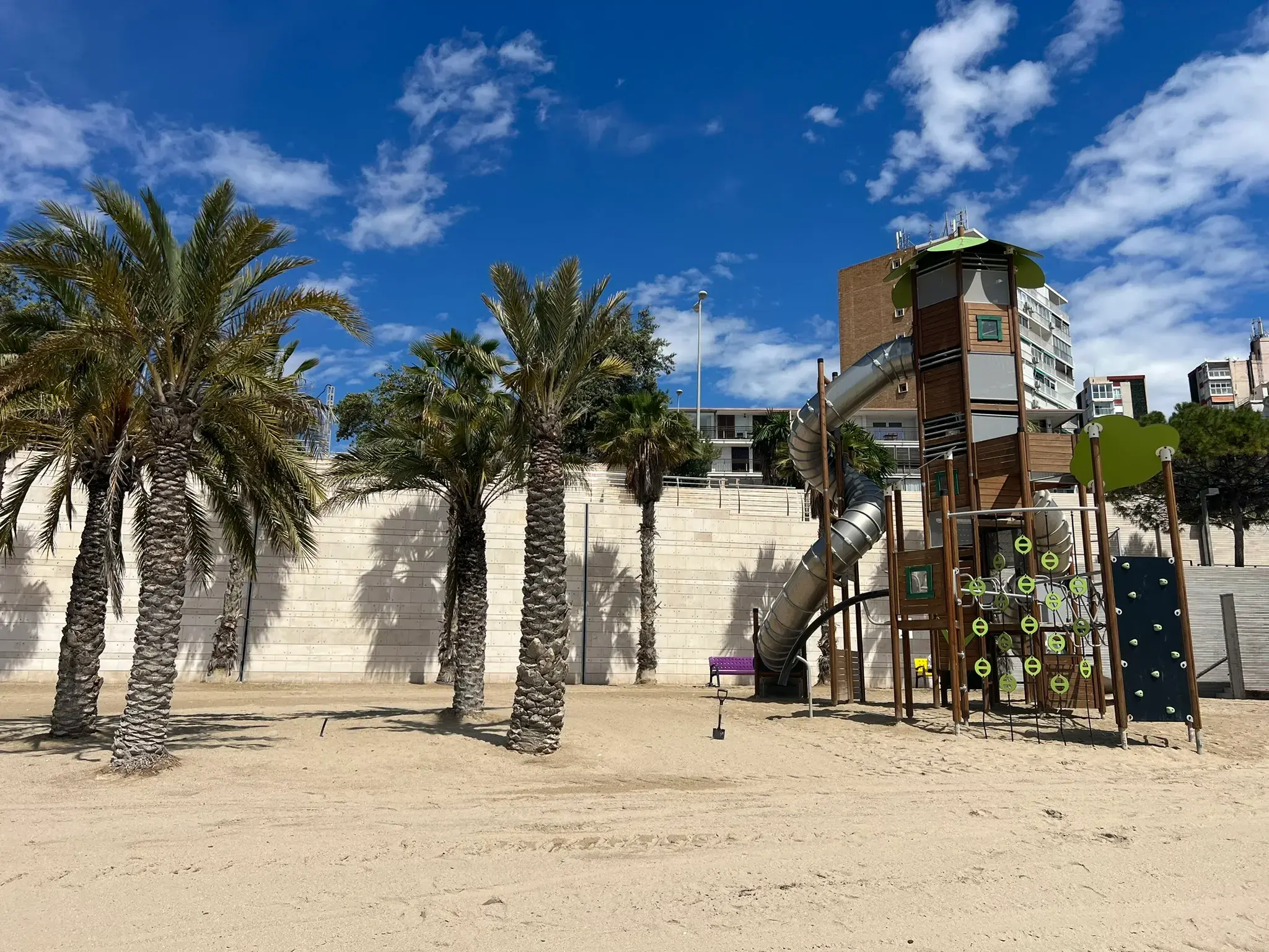 Modern and safe playground structure set right on the sand at Albufereta Beach in Alicante, with palm trees in the background and tall spiral slides next to the calm Mediterranean waters.