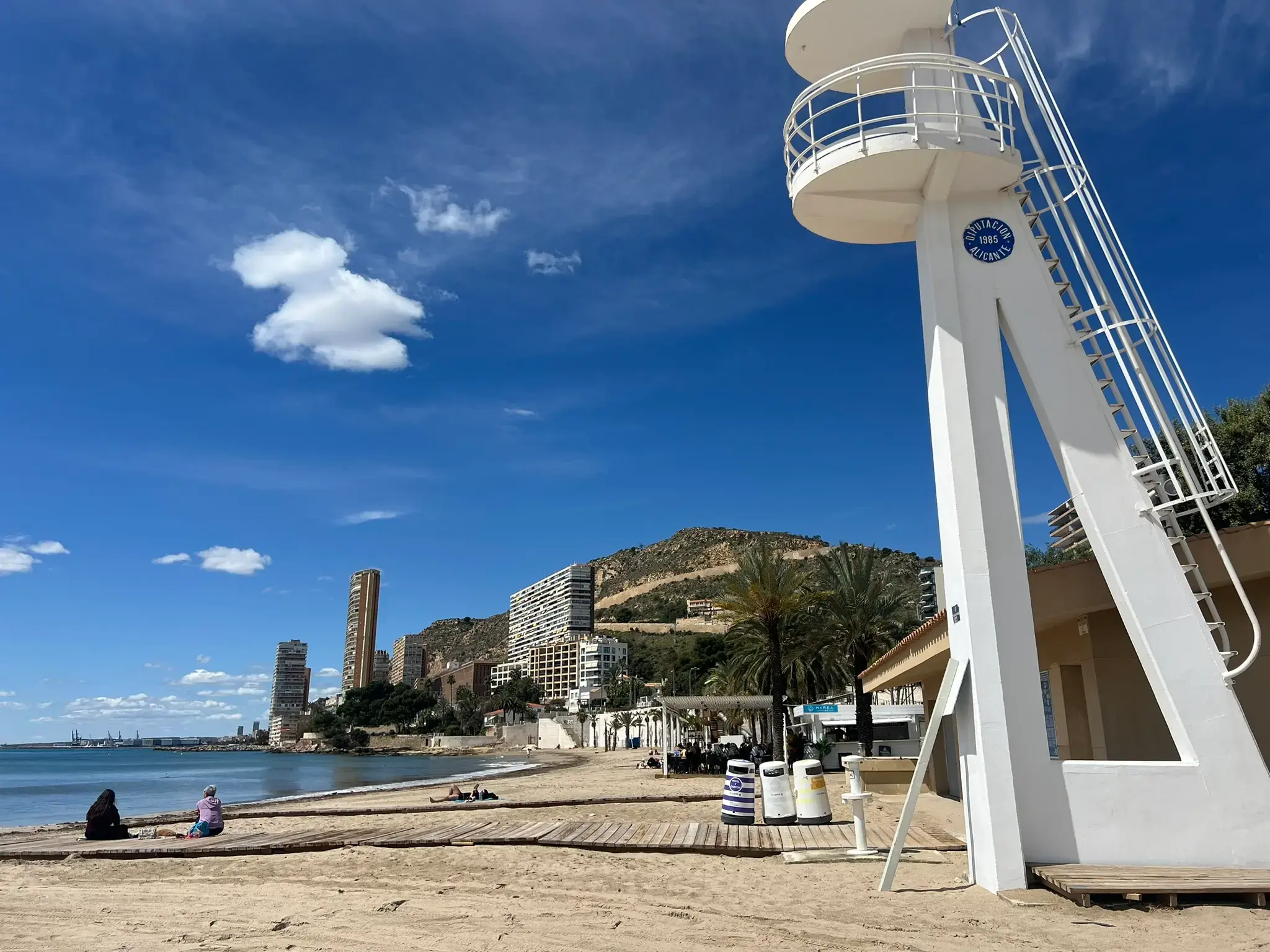 Albufereta Beach in Alicante — modern white lifeguard tower on the sandy shore, wooden walkways, clear blue Mediterranean coastline, and the Serra Grossa hill with coastal apartments.