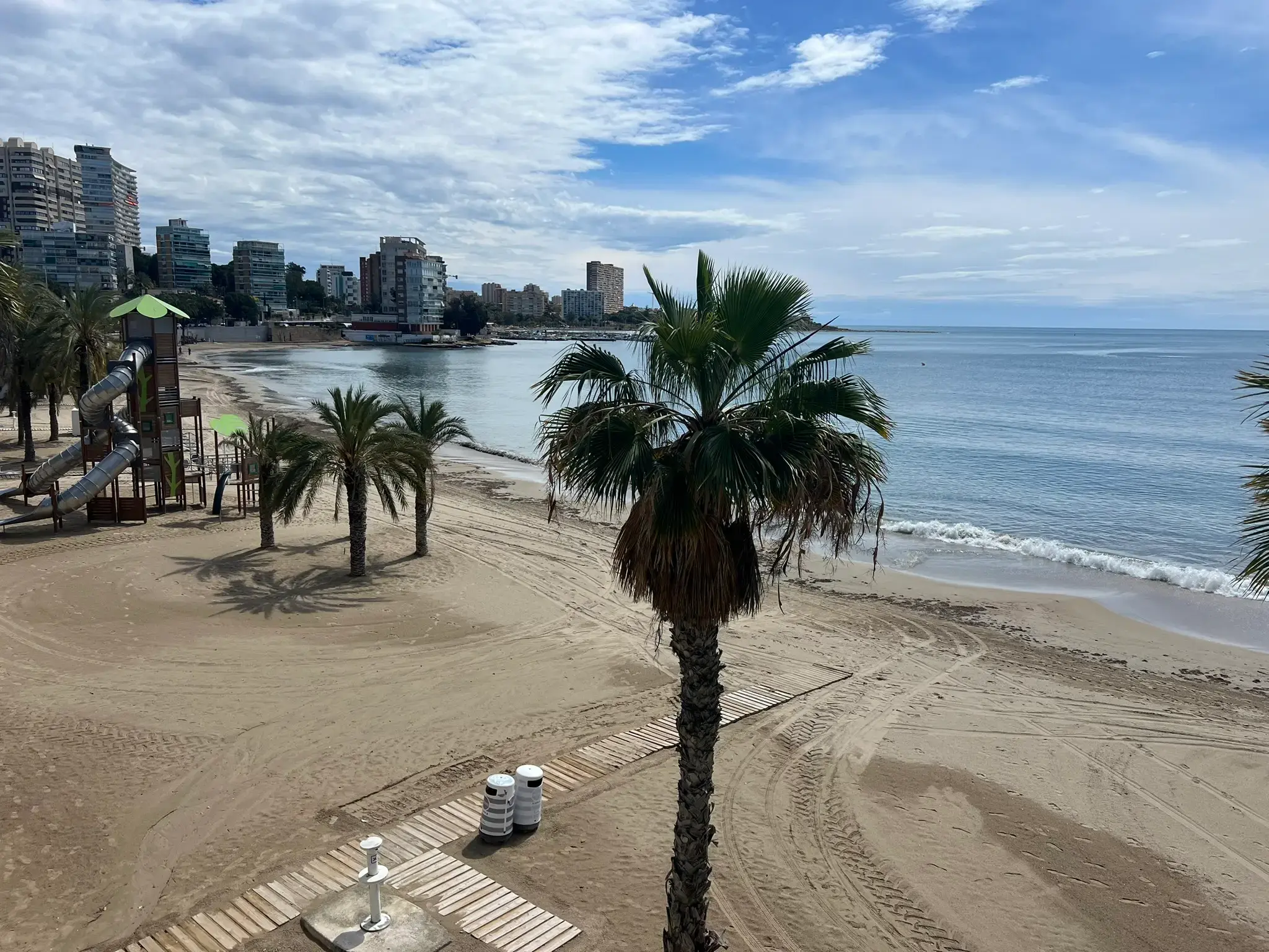 High angle view of Playa de la Albufereta featuring palm trees, a sandy beach, and a modern children's slide in Alicante, Spain.