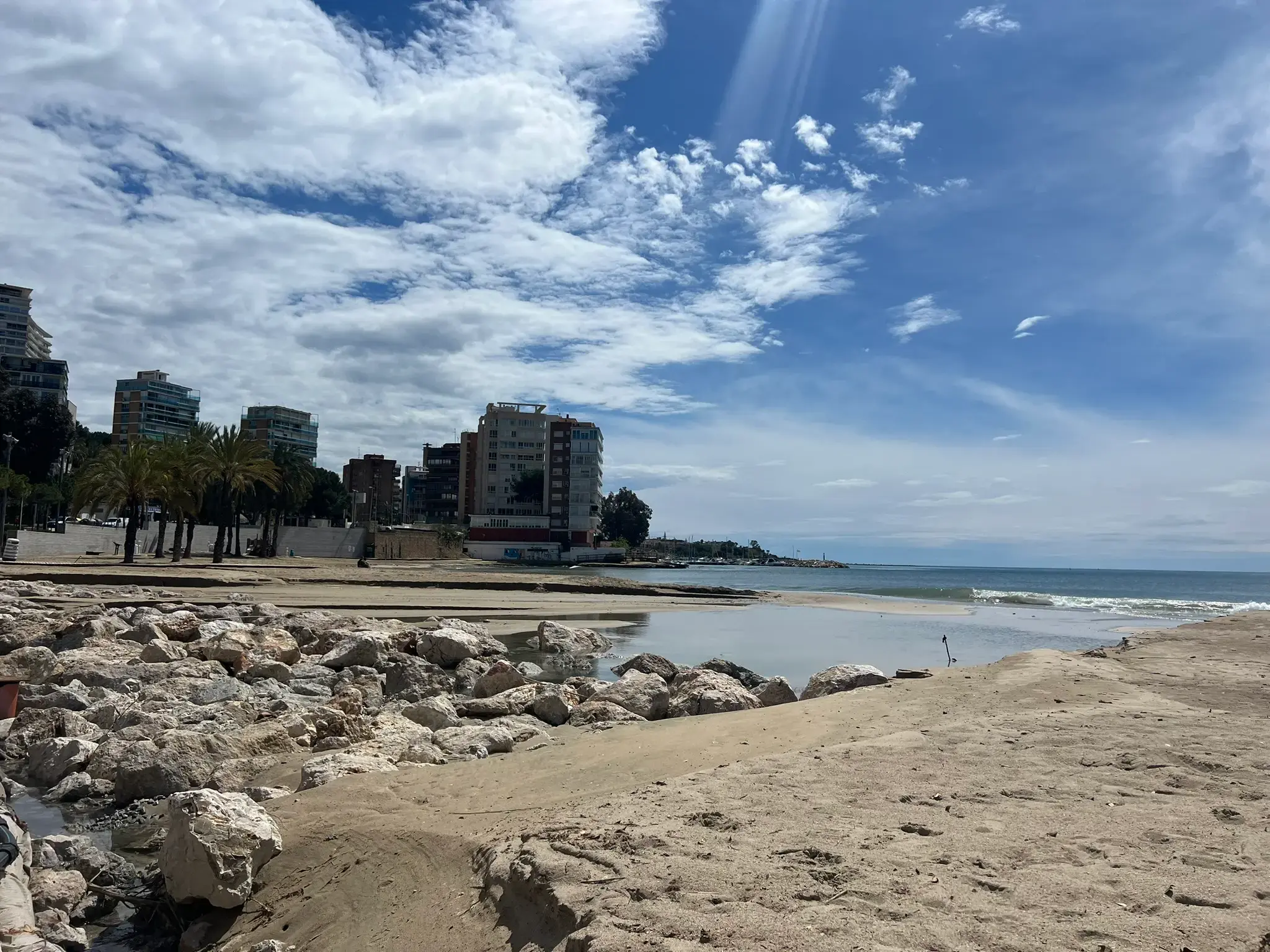 Scenic view of the coastline at Playa de la Albufereta with rocks, sand, and residential buildings under a blue sky in Alicante.