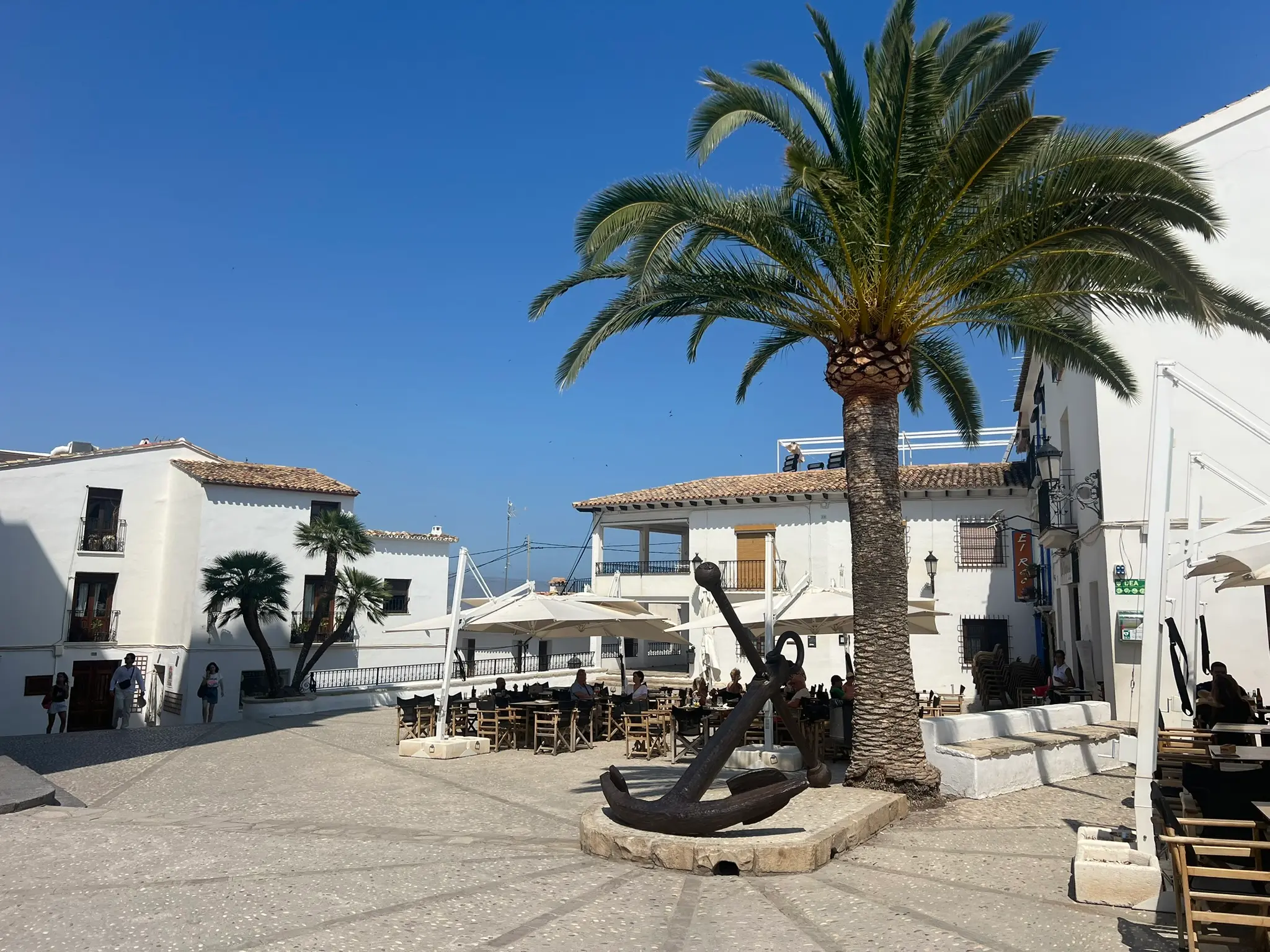 A wide-angle shot of the historic church square, Plaza de la Iglesia, in Altea old town, highlighting a massive vintage ship anchor on a stone pedestal next to a tall palm tree, with outdoor restaurant terraces and whitewashed buildings under a clear blue sky.