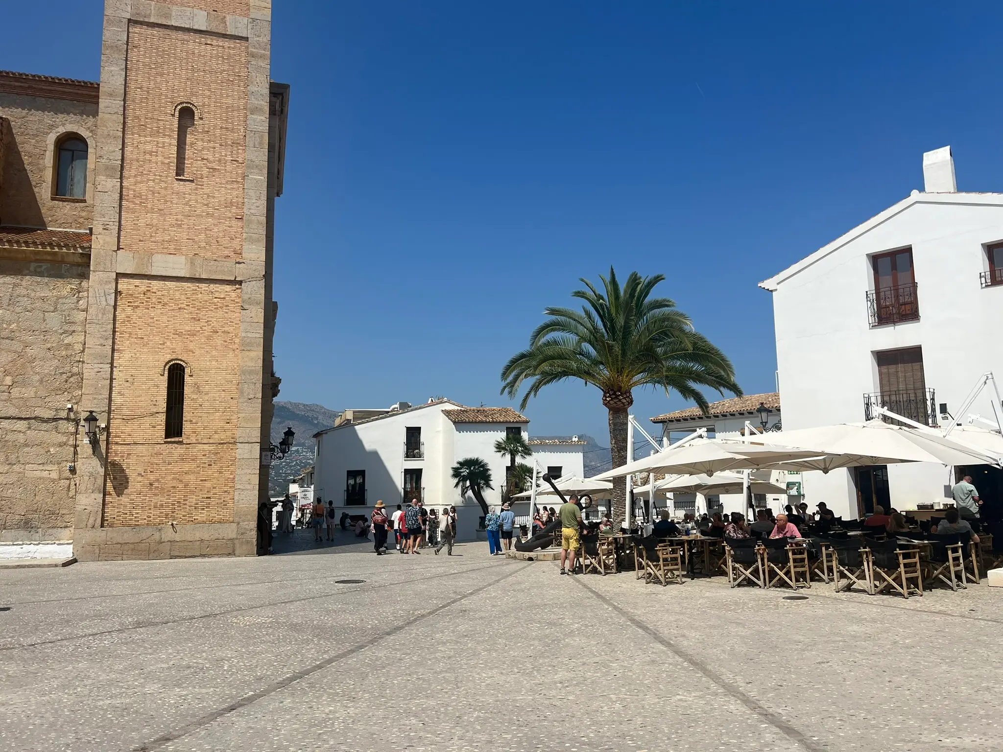 The main church square, Plaza de la Iglesia, in Altea old town, Spain, showcasing the stone bell tower of the Consuelo church, a large central palm tree, and crowded outdoor restaurant terraces under white sun umbrellas under a clear blue sky.