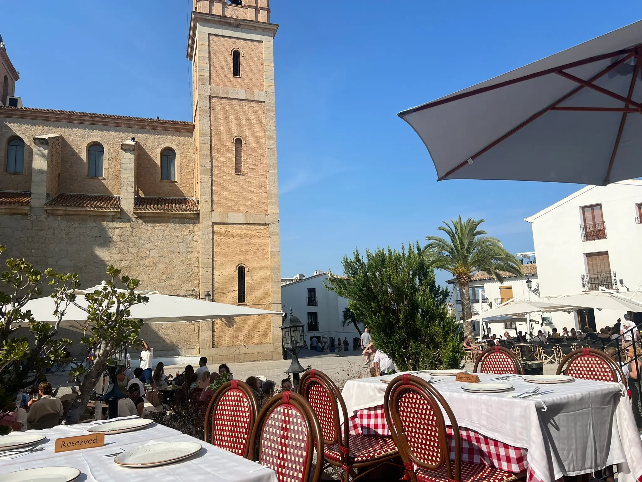 An outdoor restaurant terrace on the lively Plaza de la Iglesia in Altea, Spain. Tables are set with white tablecloths, plates, and 'Reserved' signs, surrounded by red and white woven chairs. The historic tower of the Consuelo church and a tall palm tree stand in the background under a clear sky.