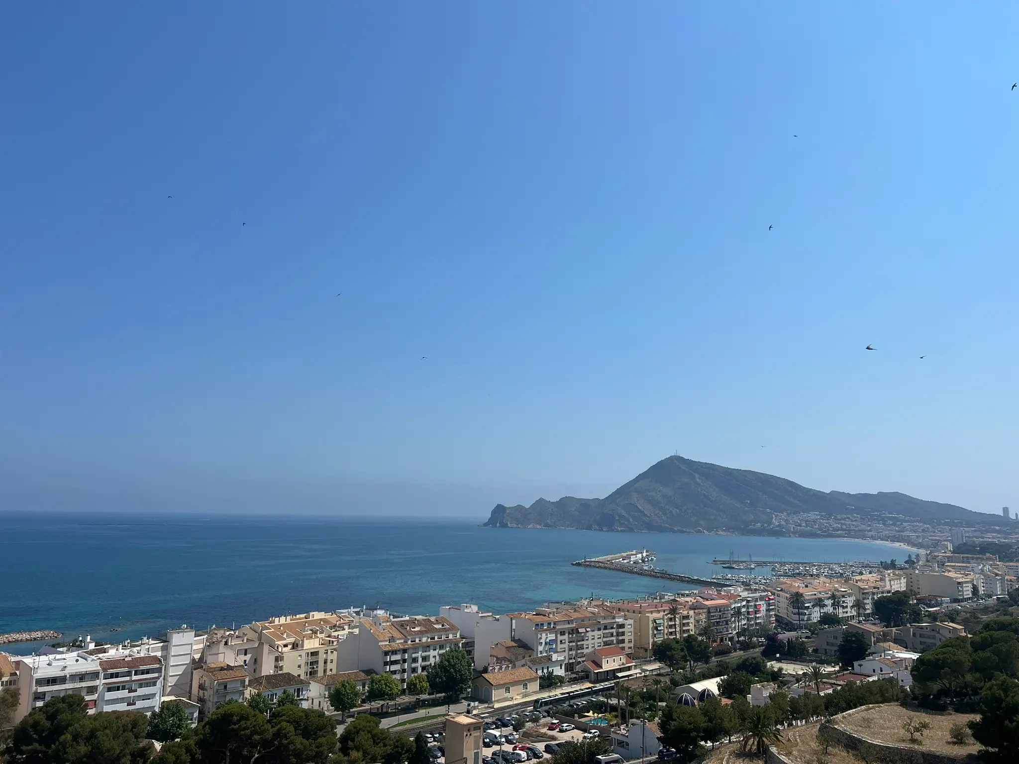 A breathtaking high-angle panoramic view of Altea coast and the Mediterranean Sea bay, showcasing the coastal town buildings, the marina port with boats, and the majestic Sierra Helada mountain cape in the background under a clear blue sky.