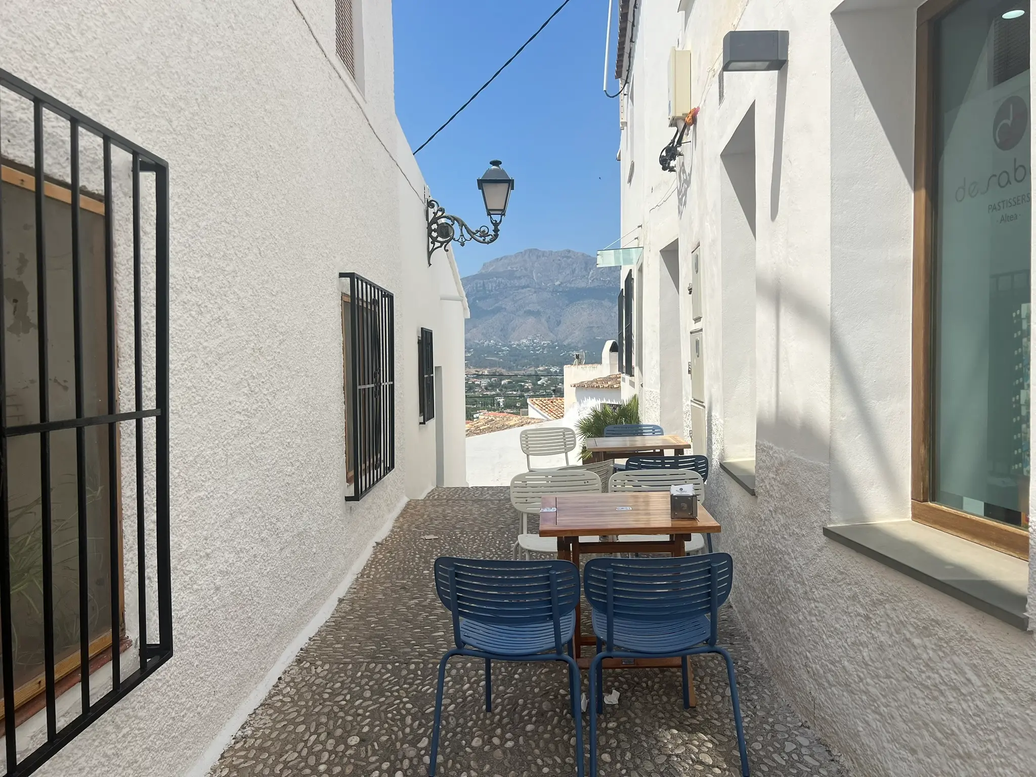 An outdoor cafe terrace in a narrow cobblestone alley of Altea old town, featuring wooden tables with blue and white chairs set against whitewashed walls with a vintage street lamp, overlooking a dramatic mountain landscape under a clear blue sky.