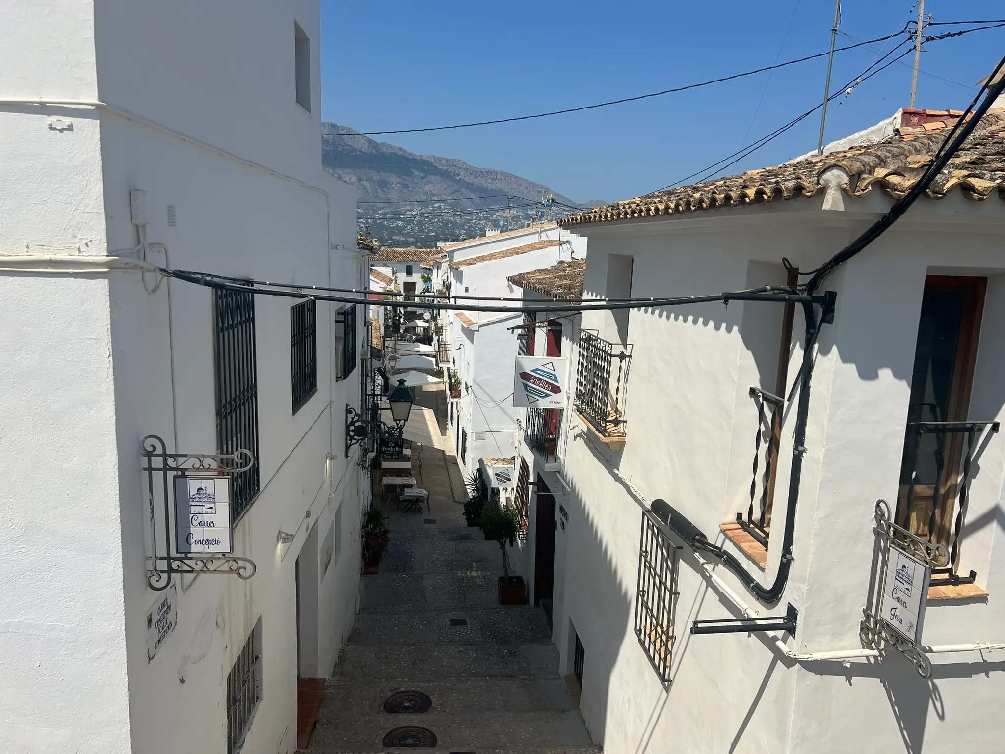 A high-angle perspective looking down a narrow cobblestone street, Carrer Concepció, in the old town of Altea, Spain. The view is framed by traditional whitewashed houses with vintage metal signs, leading to a restaurant alley with mountains in the background under a clear sky.