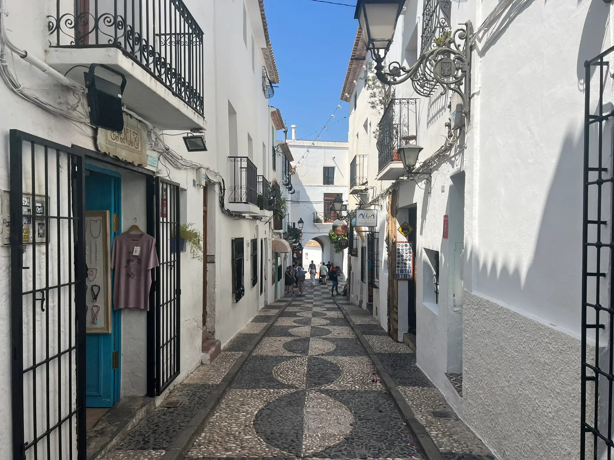 A picturesque view down Carrer Major street in Altea old town, Spain, leading towards the historic Portal Viejo stone arch. The cobblestone street features a beautiful black-and-white pebble pattern, lined with whitewashed buildings, artisan souvenir shops, and traditional wrought-iron balconies under a blue sky.