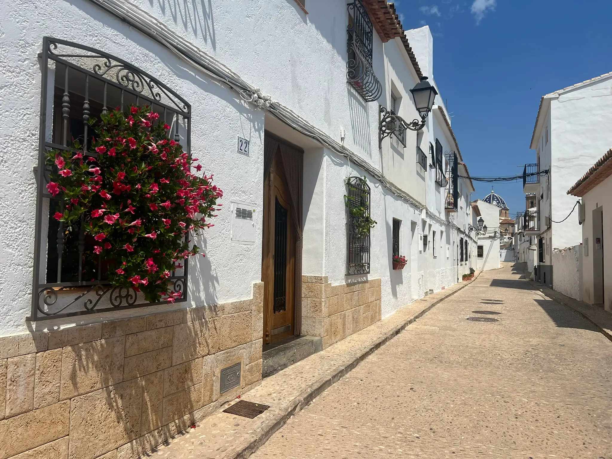 A beautiful cobblestone street in Altea old town featuring a whitewashed house with vibrant pink flowers on a wrought-iron window grille, a traditional wooden door, and the iconic blue-and-white tiled church dome visible in the background under a bright blue sky.
