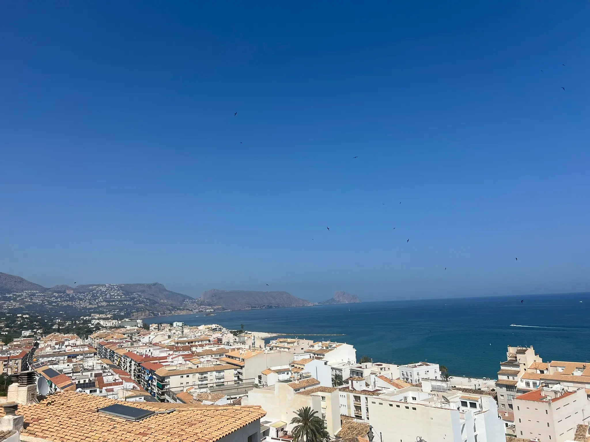 A sweeping panoramic view from the old town of Altea, Spain, overlooking white coastal buildings with terracotta tiled roofs, the Mediterranean Sea, and the distant iconic rock of Peñón de Ifach in Calpe under a vast clear blue sky.