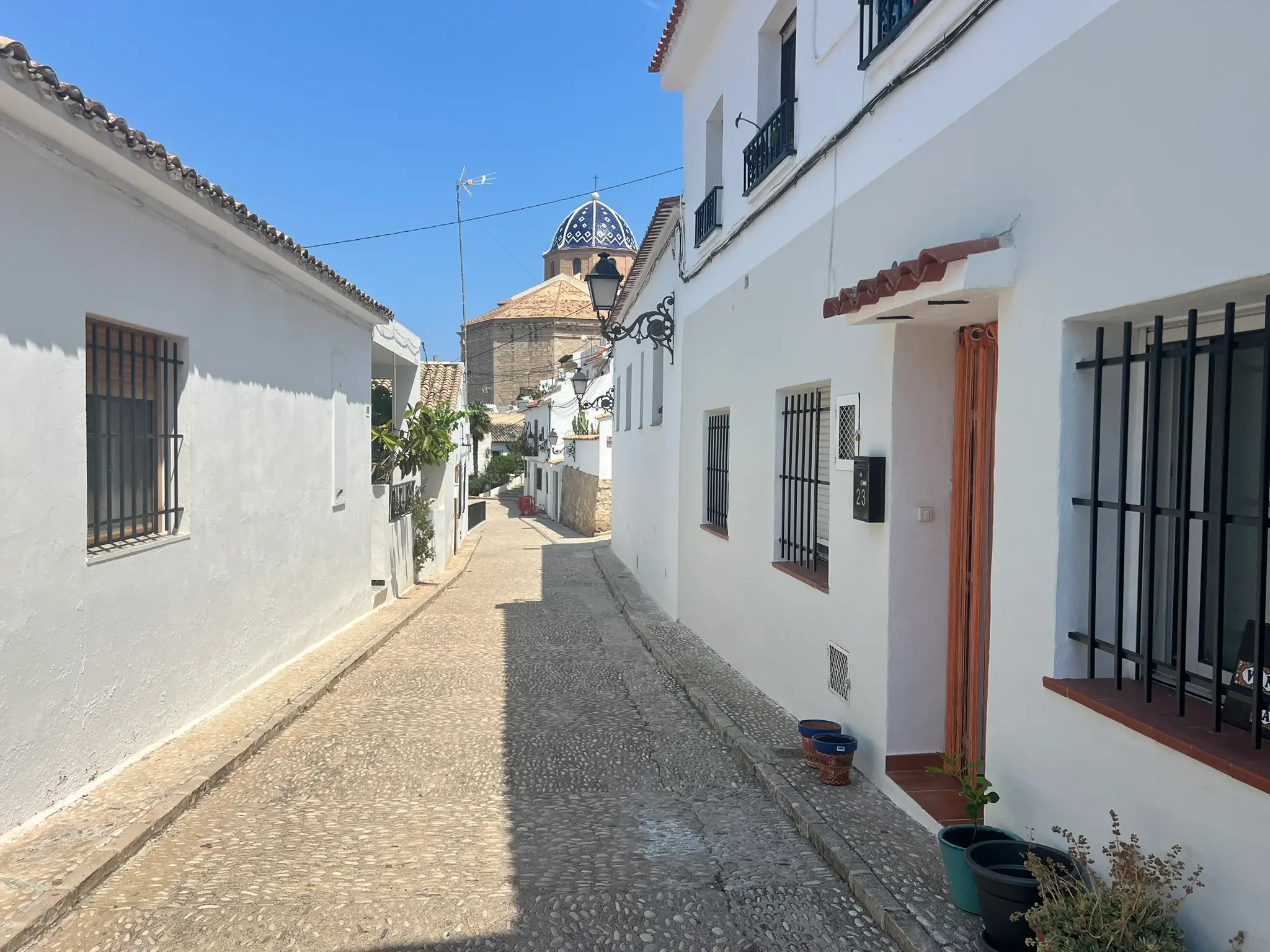 A perspective view of a traditional cobblestone street in Altea old town, lined with whitewashed houses and vintage street lamps, leading directly toward the iconic blue-and-white tiled church dome under a clear blue sky.