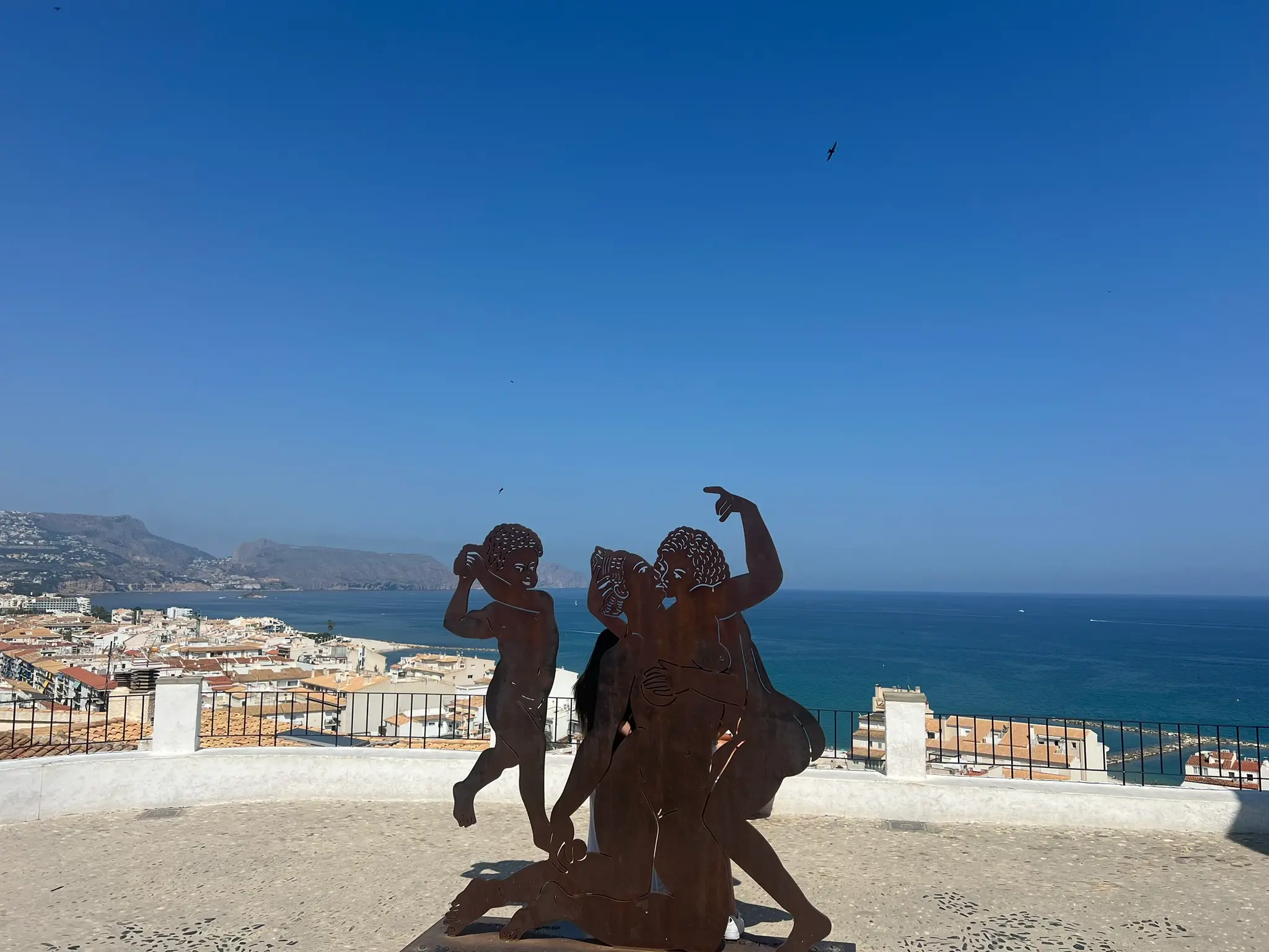 A metal silhouette sculpture of a family by artist Toni Marí stands on a scenic viewpoint overlooking the Mediterranean Sea and the coastal town of Altea, Spain, under a clear blue sky.