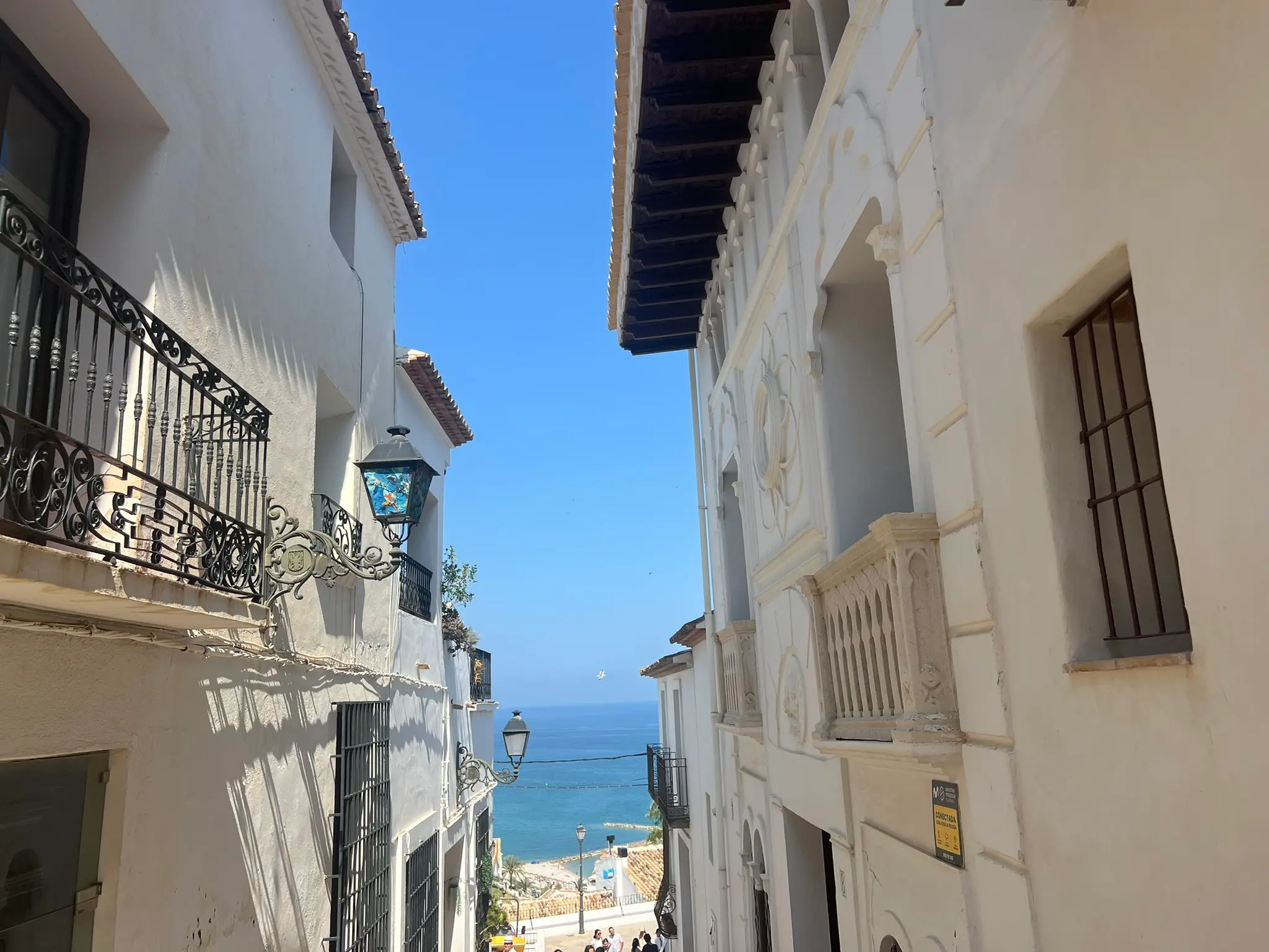 A high-angle perspective of a quiet, sunlit cobblestone street sloping down in Altea old town, lined with traditional whitewashed houses, vintage black street lamps, and green decorative plants, revealing a glimpse of the Mediterranean Sea in the background under a clear blue sky.