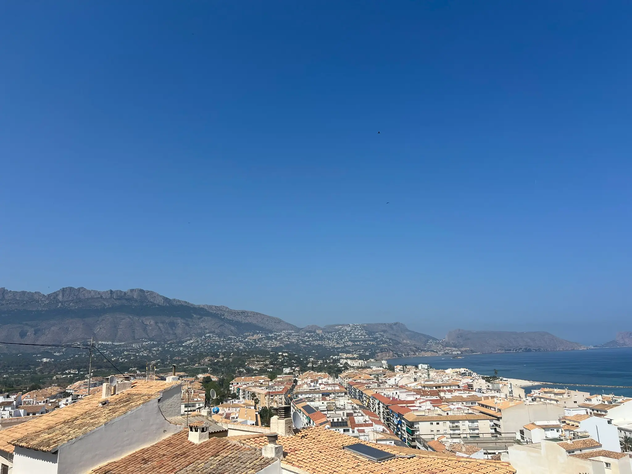 A wide panoramic overlook from Altea old town, Spain, showing traditional terracotta tiled rooftops, the dense coastal town layout, the majestic Sierra Bernia mountain range in the background, and the blue Mediterranean Sea under a clear sky.