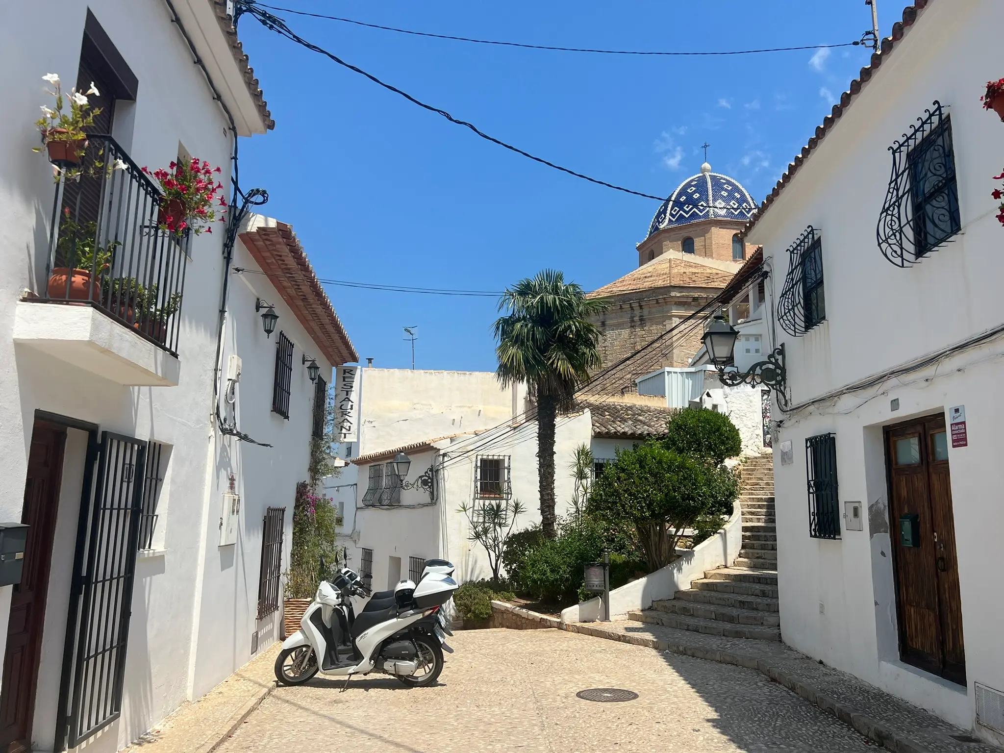 A sunny street in the old town of Altea, Spain, featuring a white scooter parked on a cobblestone road, a green palm tree, and stone steps leading up to the iconic blue-and-white tiled church dome, surrounded by traditional white houses under a clear blue sky.