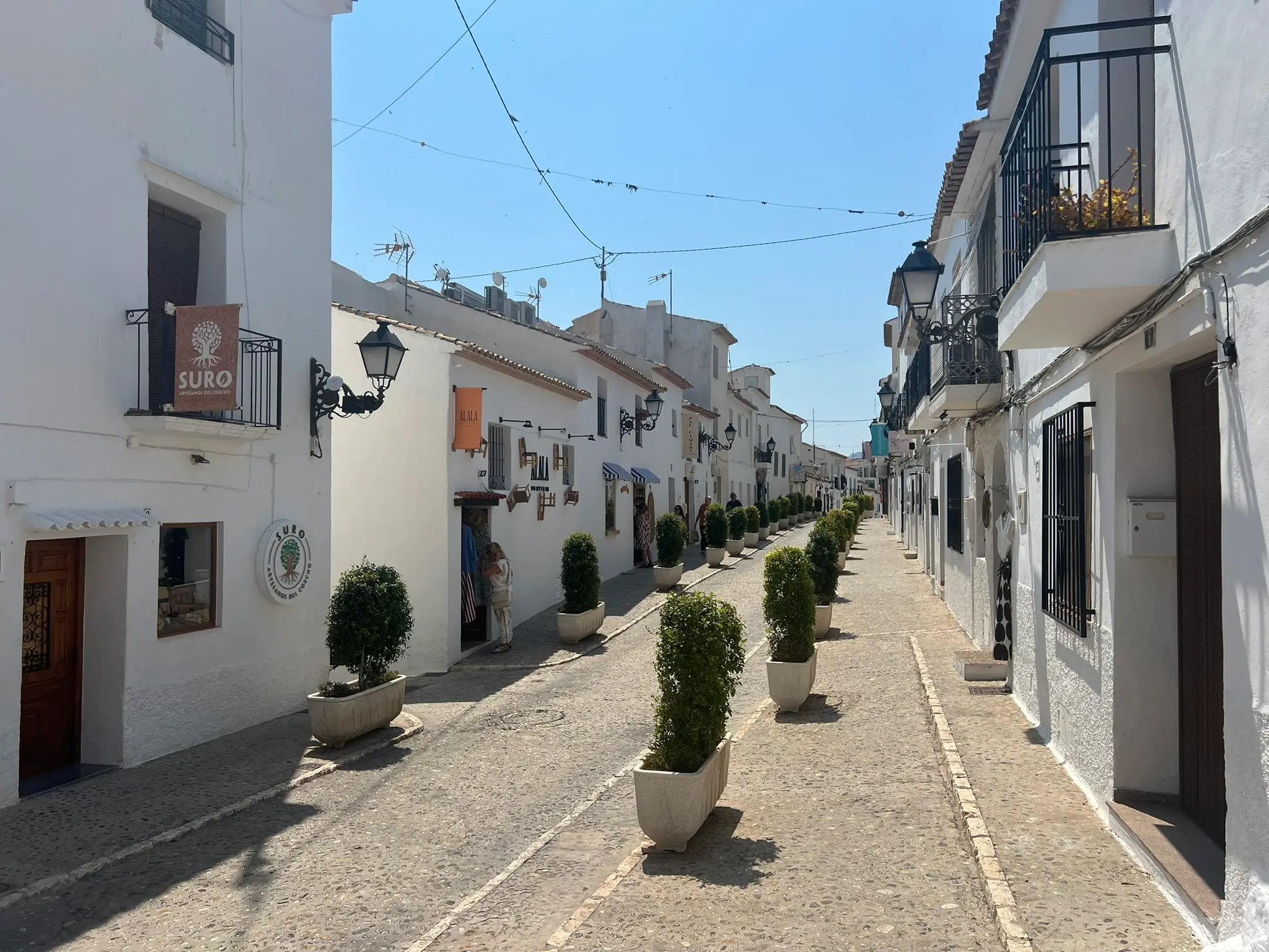 A scenic perspective of a sunny shopping street in the old town of Altea, Spain, lined with traditional whitewashed buildings housing local craft boutiques and shops, with a neat row of green potted trees dividing the cobblestone pavement under a bright blue sky.
