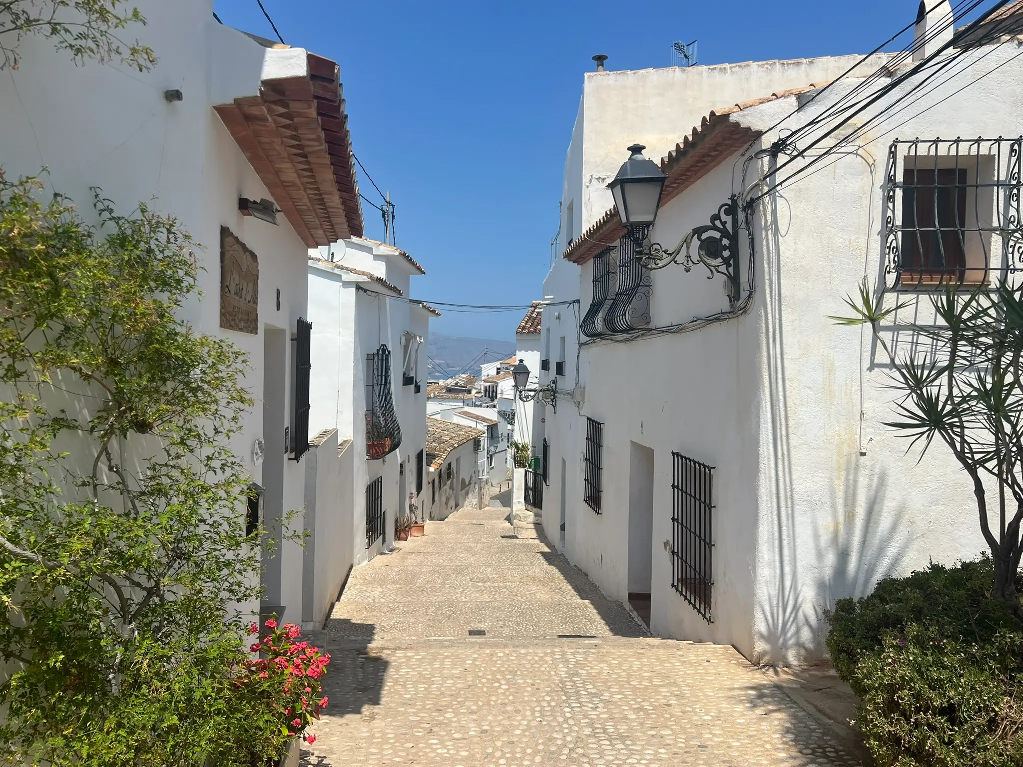 A view looking down a narrow, sunlit street in the old town of Altea, Spain. The whitewashed buildings feature traditional wrought-iron and carved stone balconies with vintage street lamps, framing a glimpse of the blue Mediterranean Sea and a distant beach at the end of the slope.