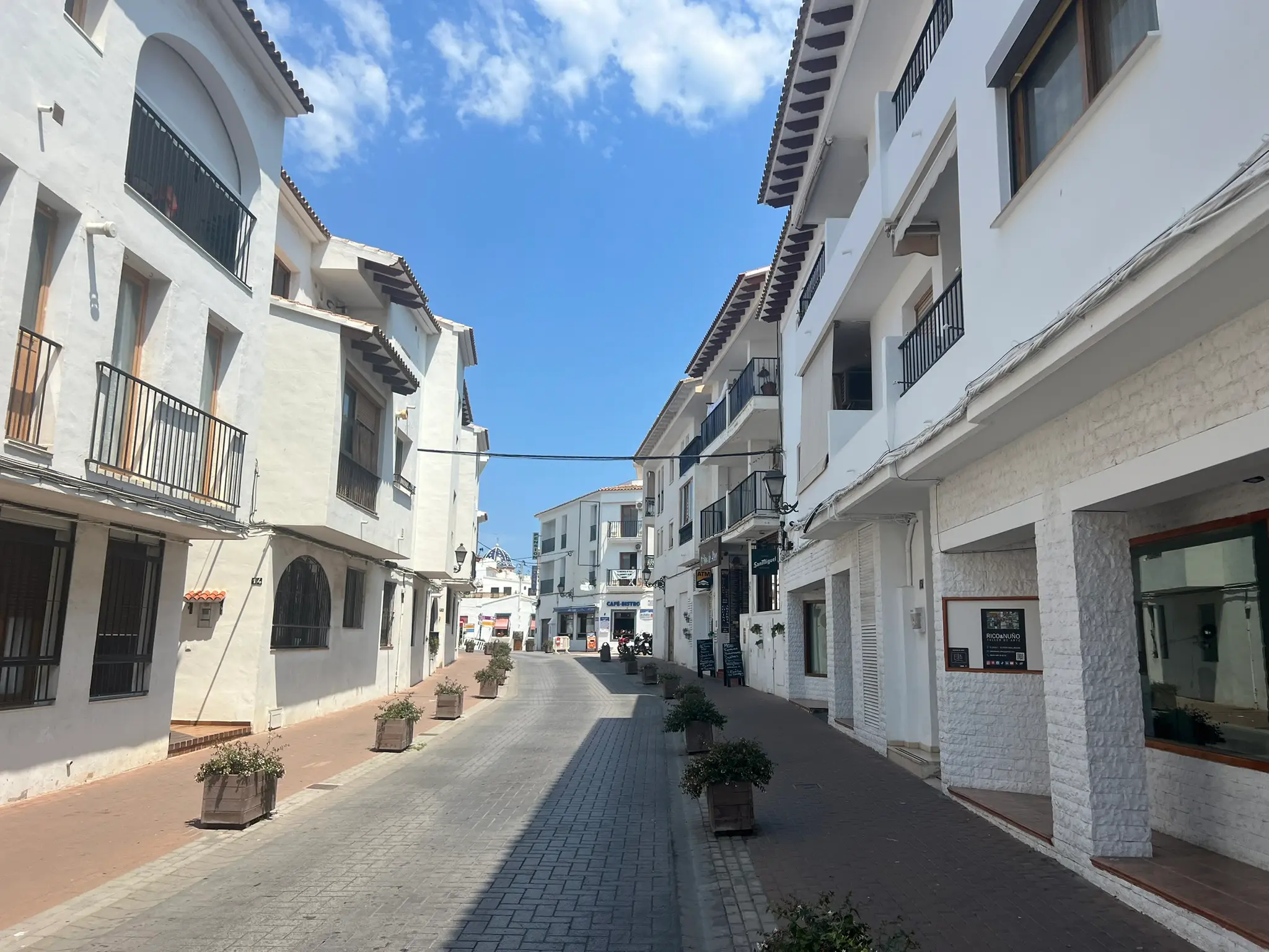 A scenic view of a narrow paved street in Altea old town (Casco Antiguo) with traditional white-washed houses under a clear blue sky, capturing the authentic Mediterranean architecture of Costa Blanca, Spain.