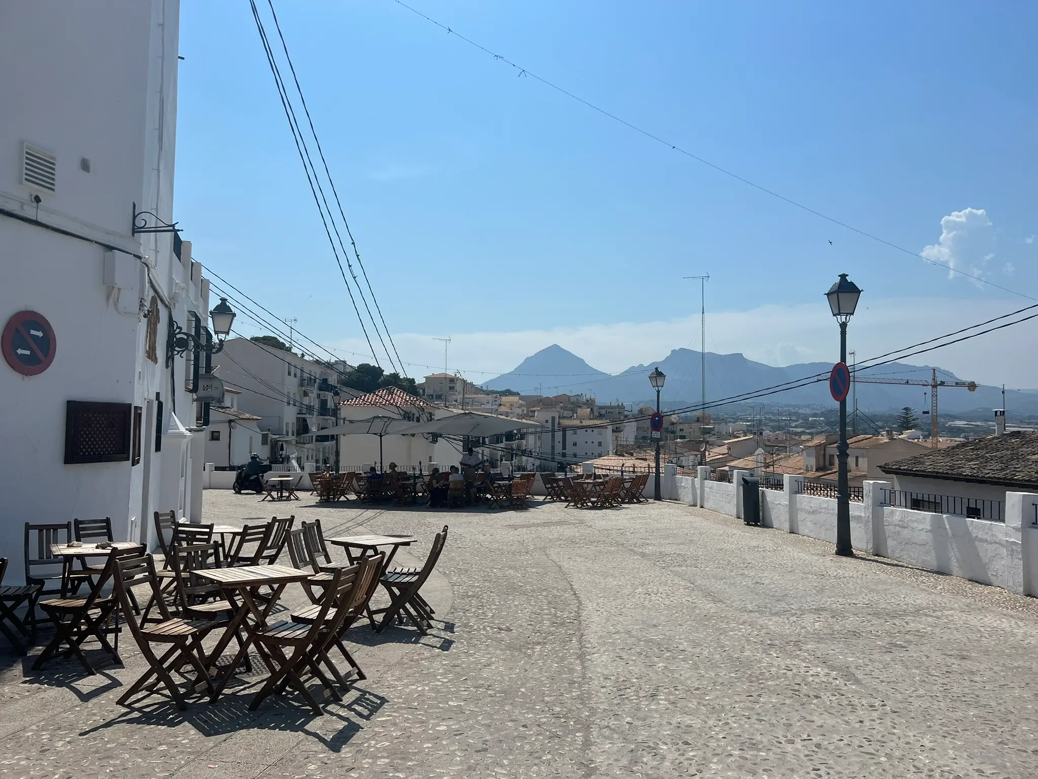 An open-air square and restaurant terrace in the old town of Altea, Spain, featuring empty wooden tables and chairs on a cobblestone pavement, vintage street lamps, and a panoramic view of distant mountains under a clear blue sky.