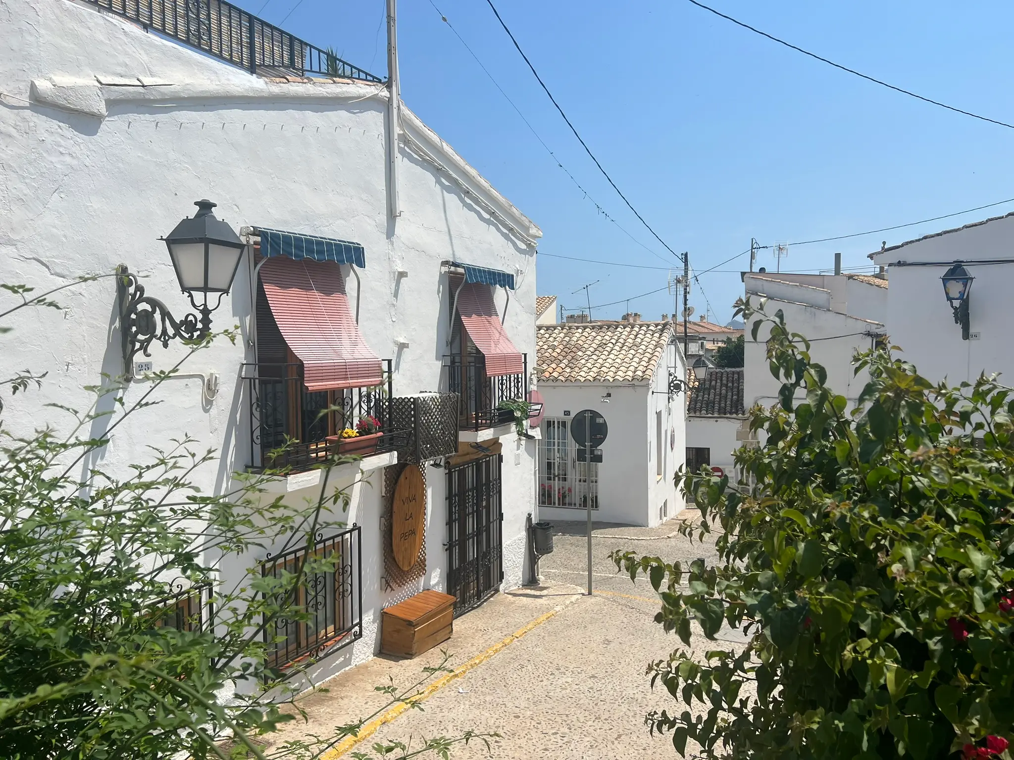 A picturesque narrow street in the old town of Altea, Spain, featuring traditional white houses with wrought-iron balconies, vintage street lamps, and pink window blinds under a clear sunny sky, framed by green Mediterranean plants.