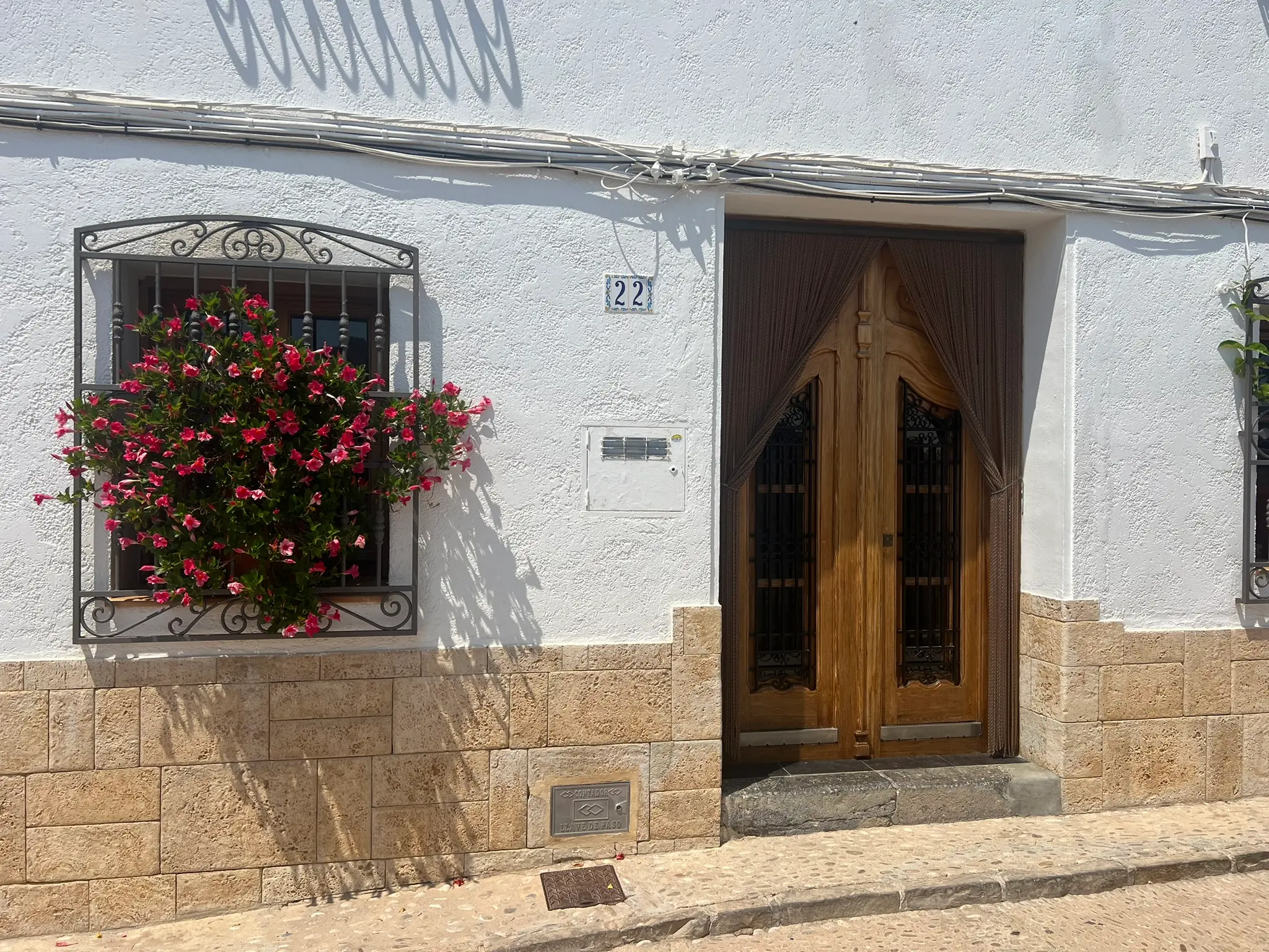 A close-up frontal view of a traditional house facade in Altea old town, numbered 22, showcasing a rustic wooden double door with a sun curtain, textured stone and whitewashed walls, and a wrought-iron window grille bursting with vibrant pink flowers.