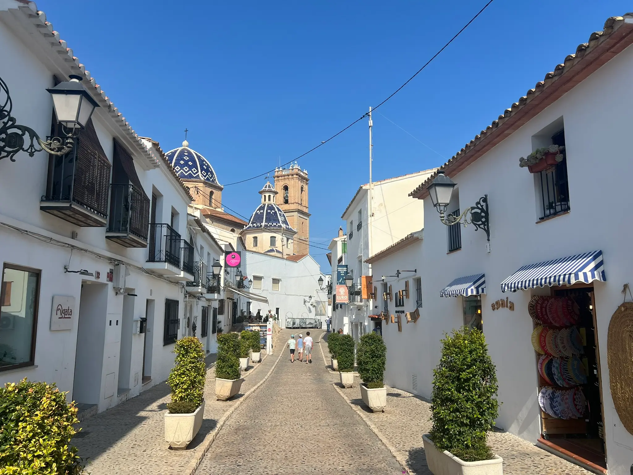 A perfectly symmetrical view looking up Calle San Miguel in Altea, Spain. The whitewashed pedestrian street features souvenir boutiques with striped awnings and potted plants, leading directly towards the iconic blue-tiled domes and bell tower of the Consuelo church under a clear sky.