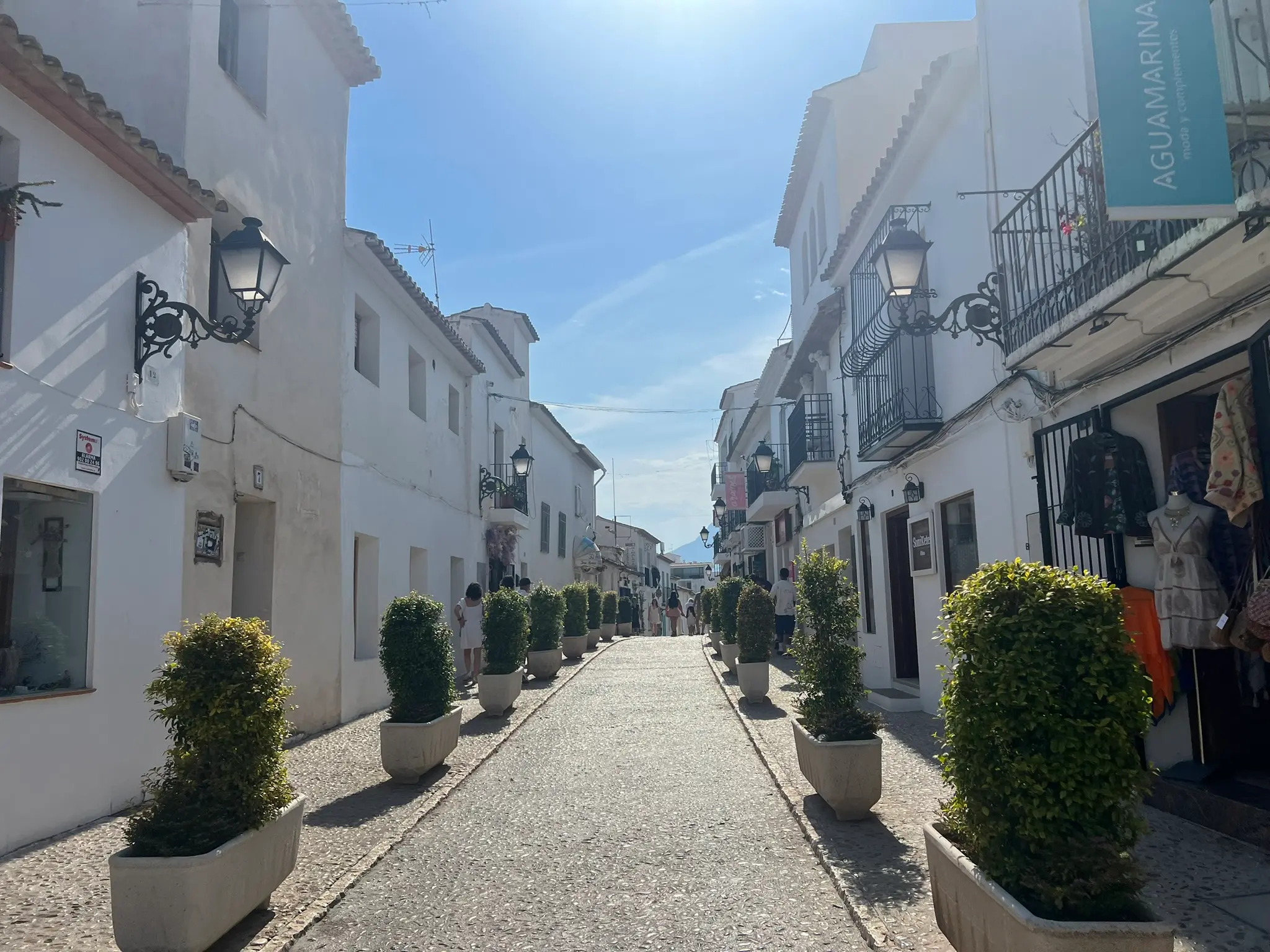 A symmetric view along the historic Calle San Miguel in Altea old town, Spain. The cobblestone pedestrian street is lined with whitewashed boutique shops, vintage black iron lanterns, and a neat row of green bushes in rectangular stone planters under a bright blue sky.