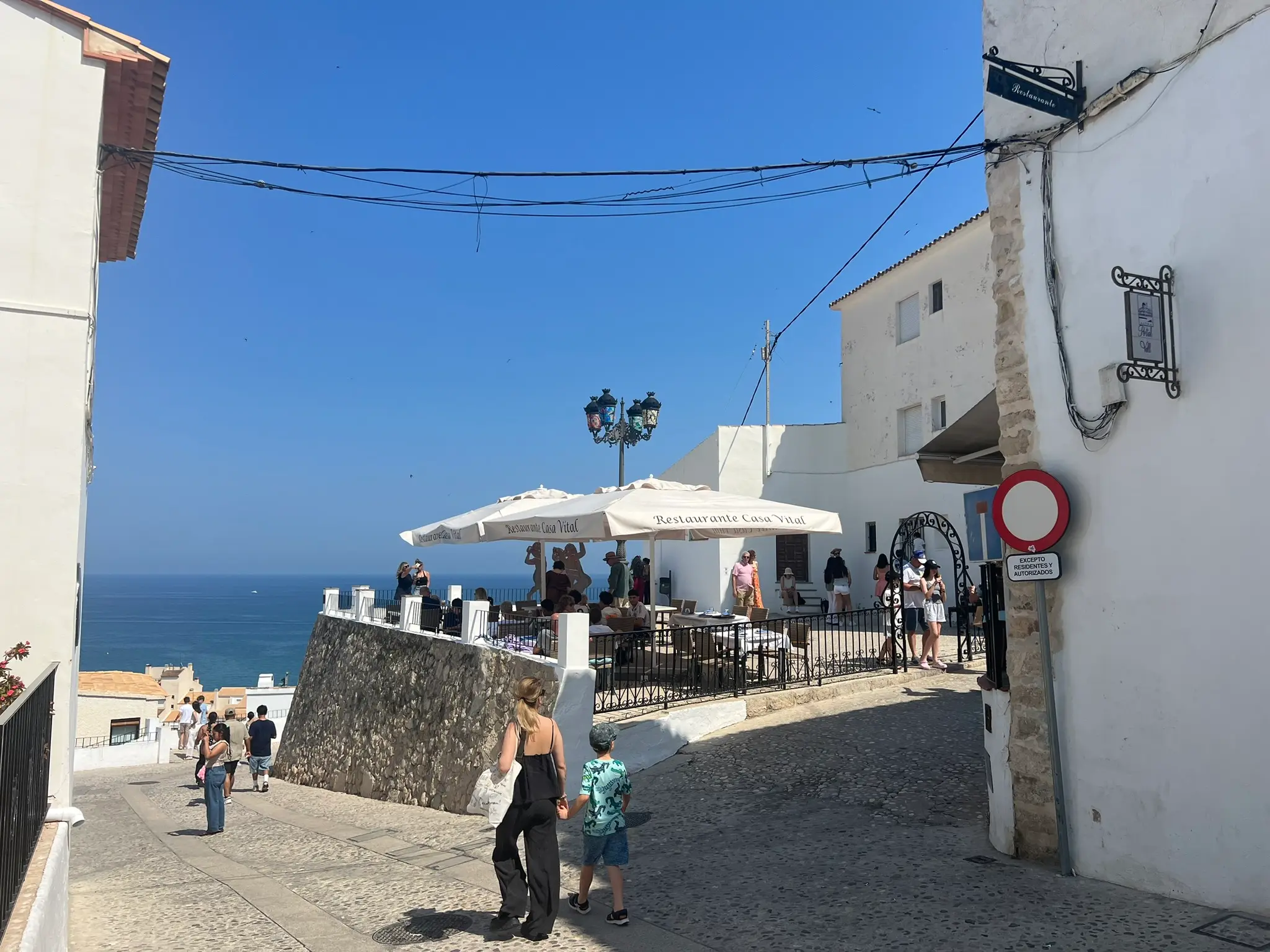 A scenic street view in the old town of Altea, Spain, showcasing the outdoor terrace of Restaurante Casa Vital with white umbrellas. Tourists walk down a cobblestone path next to a stone wall overlooking the deep blue Mediterranean Sea under a clear sky.