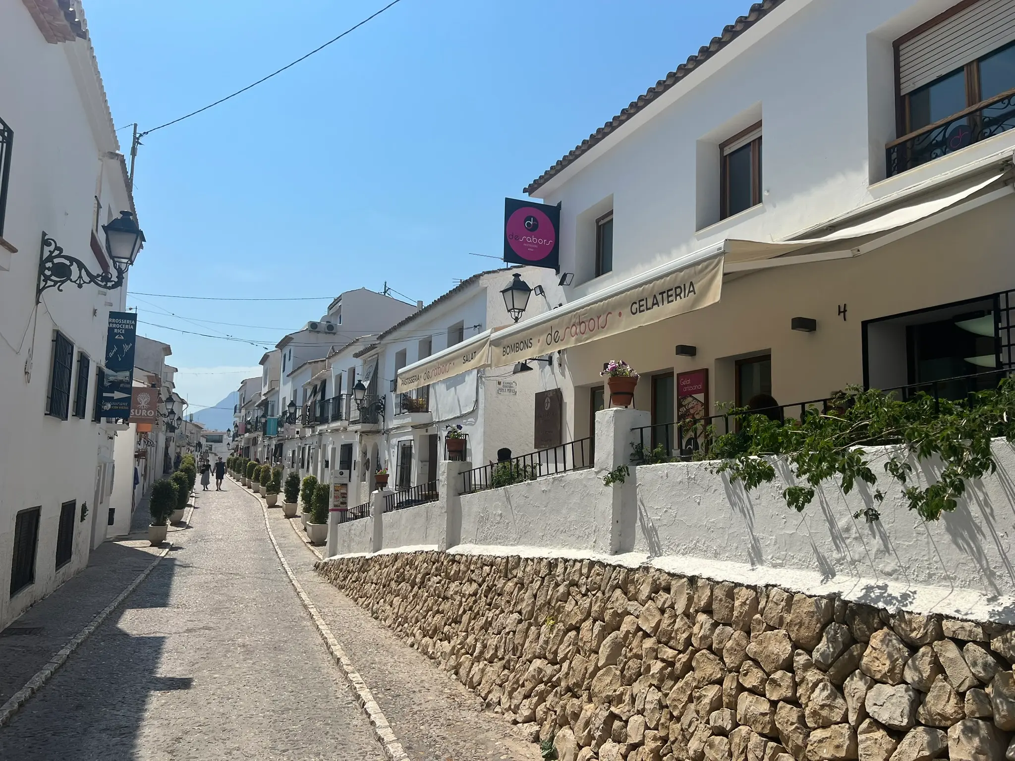 A street view in Altea old town showcasing the exterior of De Sabors Gelateria, with a cream-colored awning and a pink circular logo sign. The traditional white building features a rustic stone retaining wall on a sunlit cobblestone road under a clear sky.