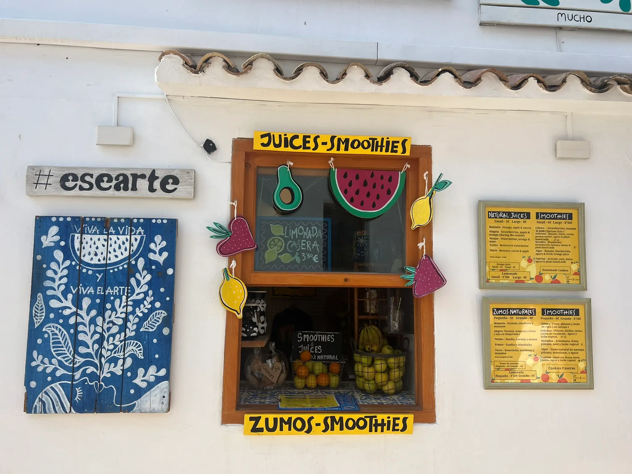 The colorful wooden window storefront of Esearte juice and smoothie bar in Altea, Spain. The display features baskets of fresh apples and oranges, hanging hand-painted wooden fruits like watermelon and avocado, framed menus in English and Spanish, and a blue painted shutter with the text 'Viva la vida y viva el arte'.