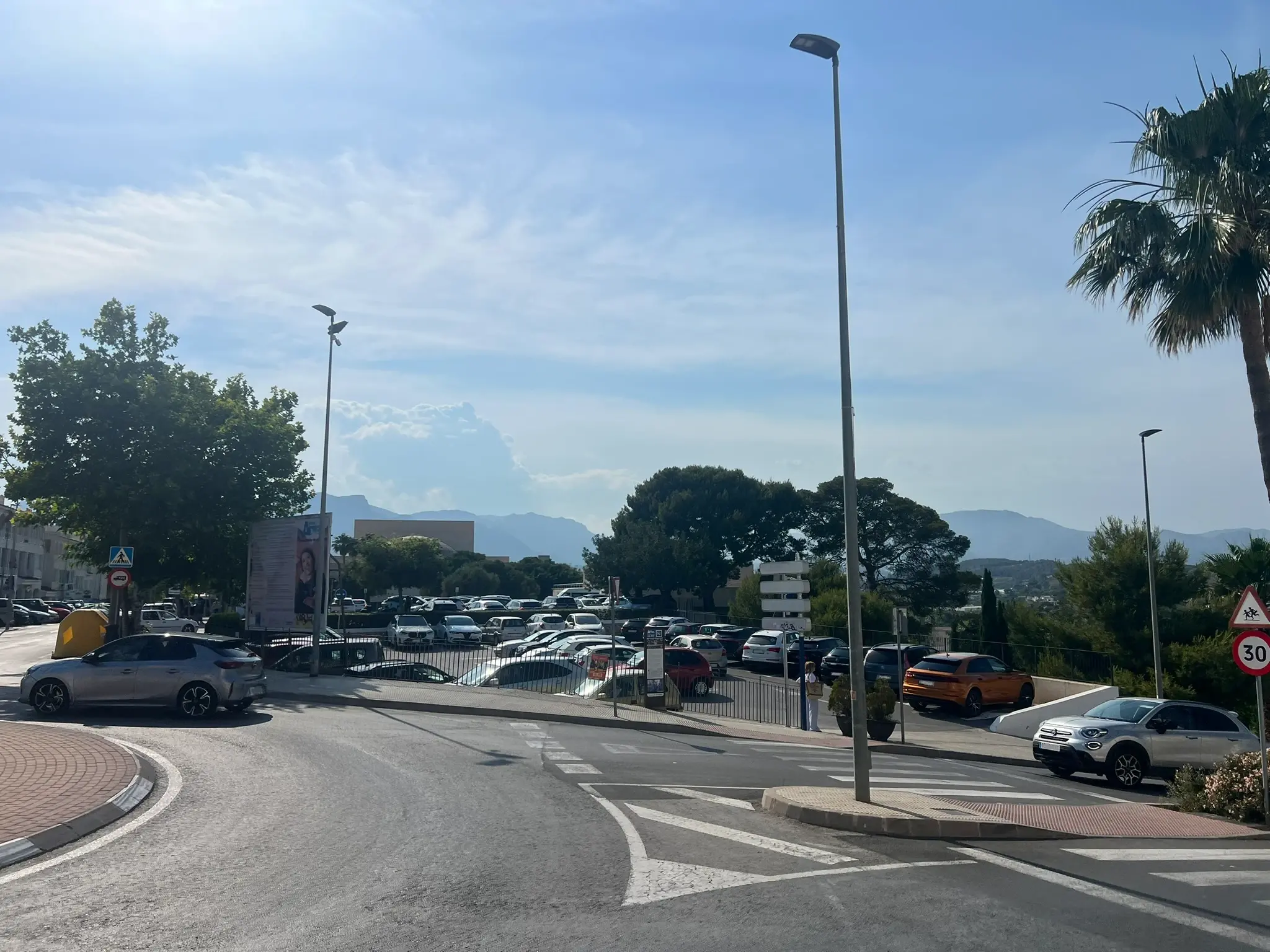 A large public outdoor parking lot packed with cars near the old town of Altea, Spain. The image shows an asphalt road intersection with a pedestrian crossing, streetlights, a palm tree, and a speed limit sign, with the mountains visible in the distance under a bright sky.