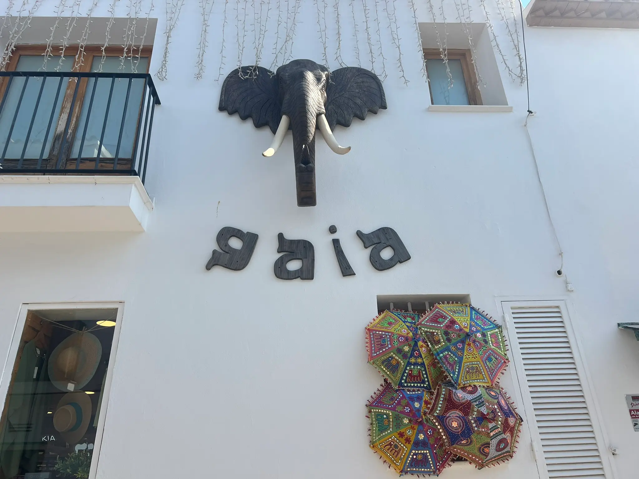 A close-up of the whitewashed facade of the Gaia boutique in Altea old town, Spain. The shop front features a large decorative wooden elephant head, the word 'gaia' in stylized dark letters, and a colorful display of embroidered traditional Indian umbrellas covering a window.