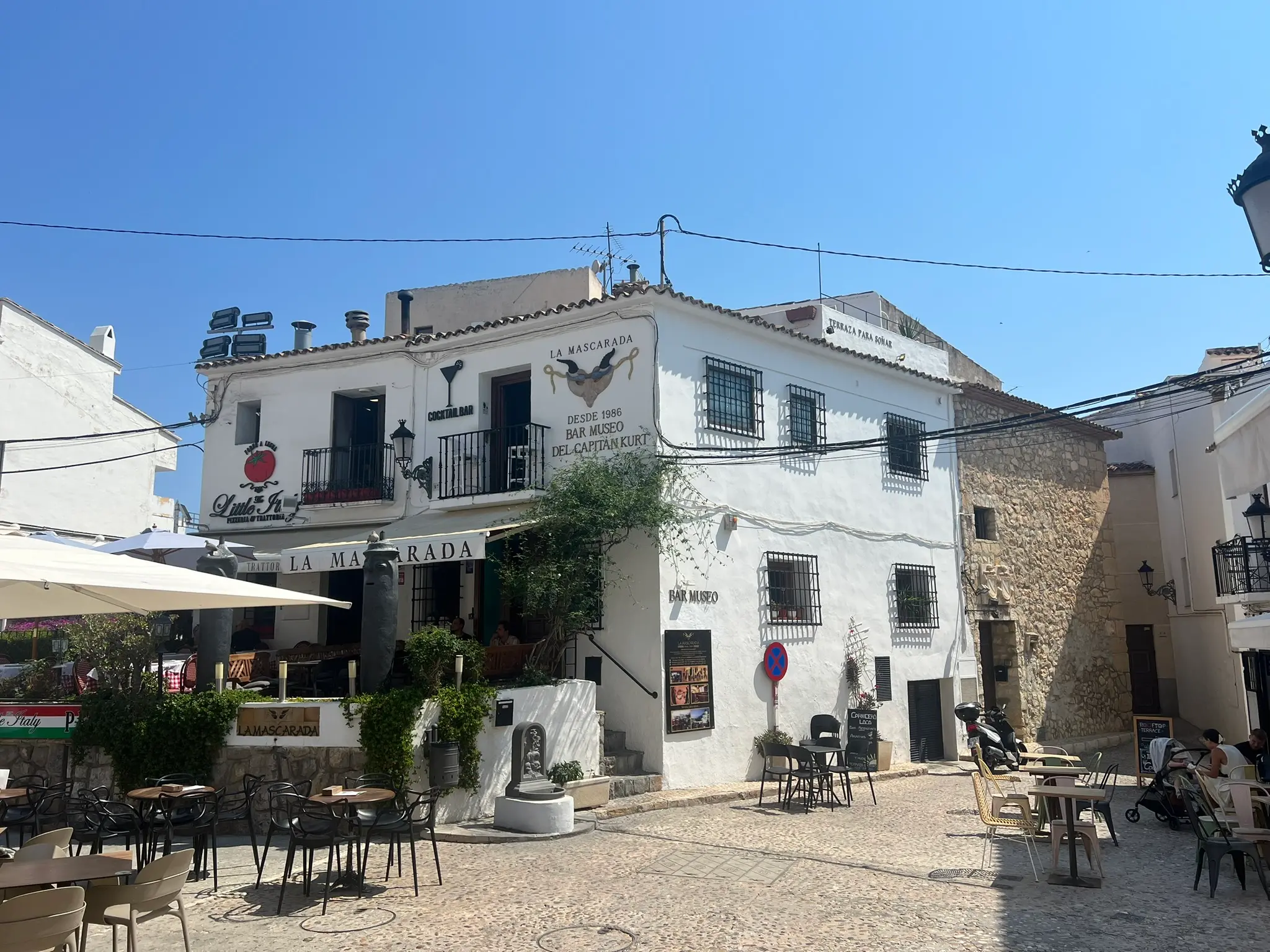 A bustling open-air square in Altea old town, featuring the whitewashed facade of La Mascarada Bar Museo, established in 1986, and Little Italy pizzeria, with outdoor cafe tables, sun umbrellas, and historical signs under a clear blue sky.