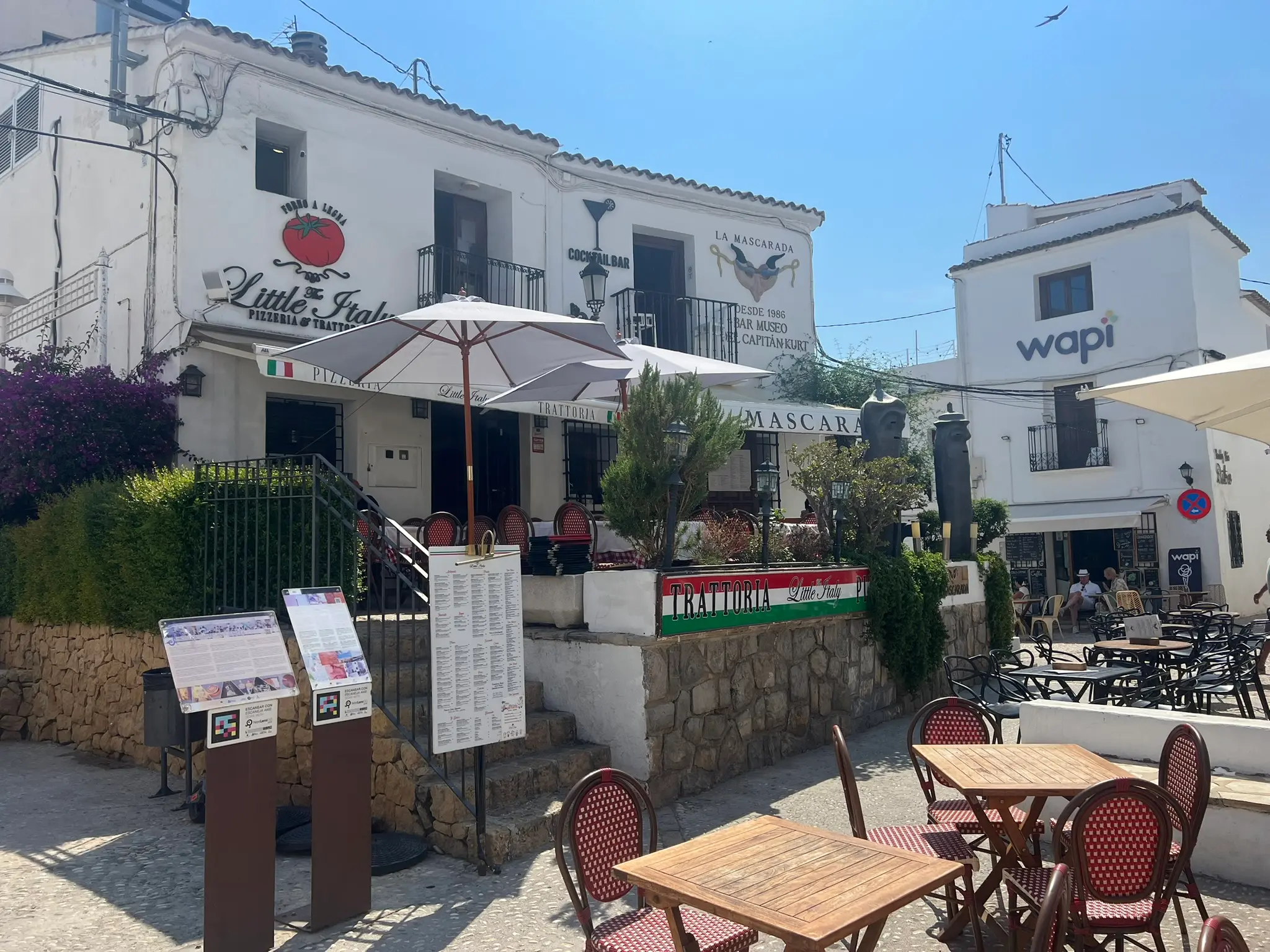 The outdoor terrace and entrance of Little Italy Pizzeria & Trattoria in Altea old town, featuring wooden tables with red-and-white chairs, large sun umbrellas, restaurant menus on display, and the whitewashed facade with a tomato logo under a bright blue sky.