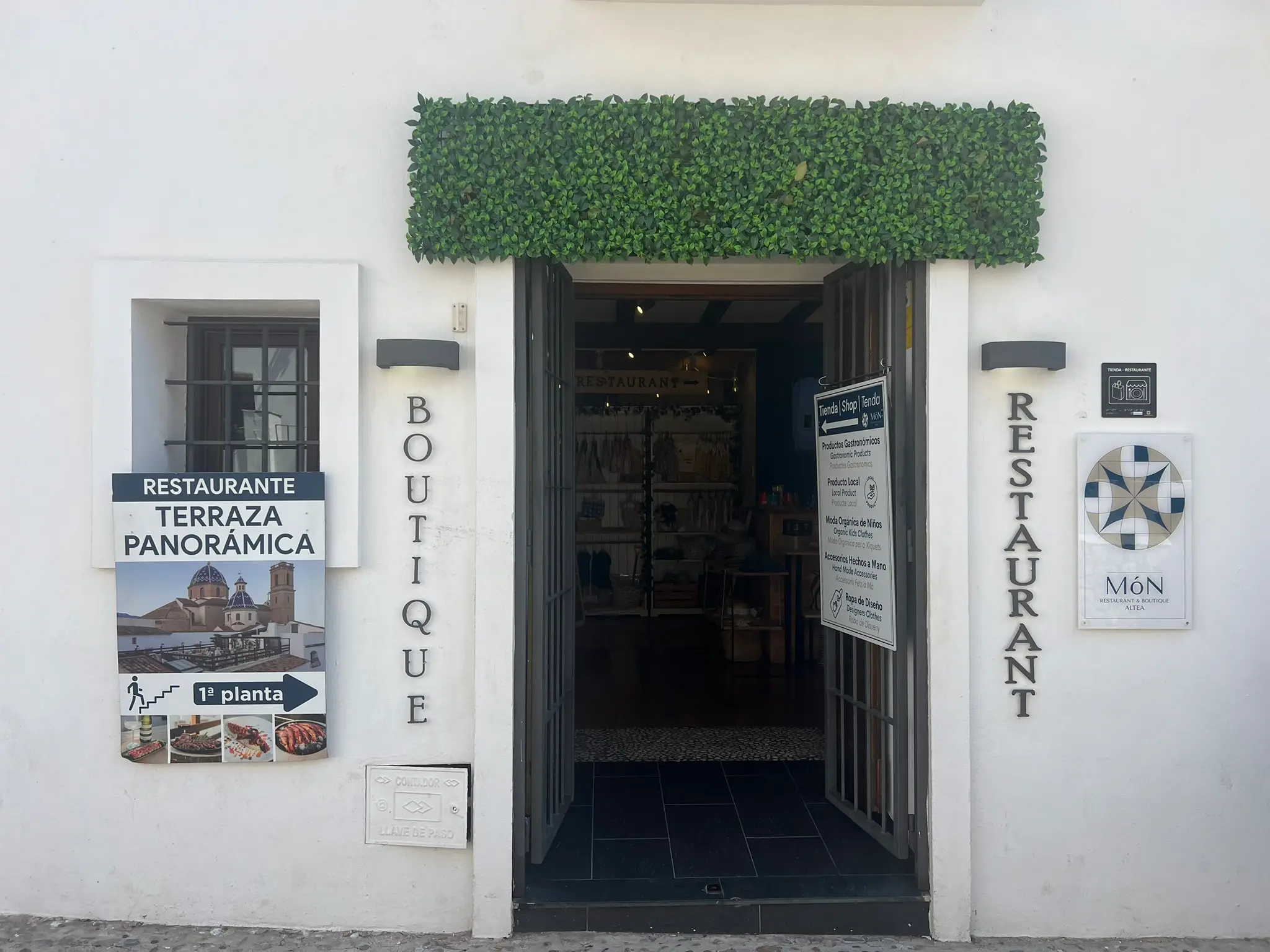 The entrance of MÓN Restaurant & Boutique in the old town of Altea, Spain, featuring white walls with 'BOUTIQUE' and 'RESTAURANT' signs, an open doorway topped with artificial green foliage, and advertising boards for a panoramic terrace overlooking the city.