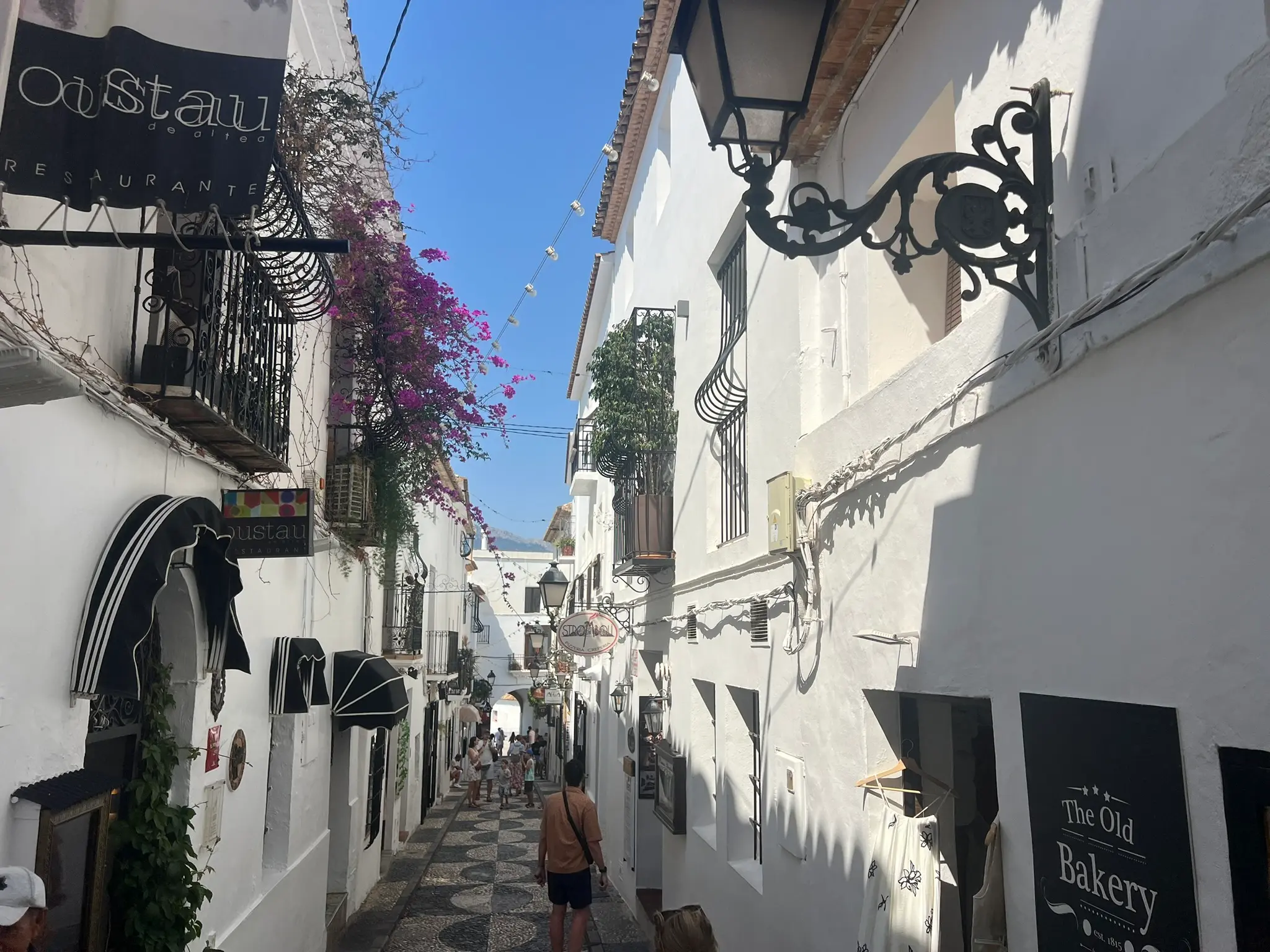 A bustling and narrow historical street, Carrer Major, in Altea old town, Spain, lined with whitewashed houses, the prominent sign of Oustau restaurant, and vibrant pink bougainvillea flowers. The scene features a decorative pebble-patterned pavement, vintage street lamps, and tourists under a clear blue sky.
