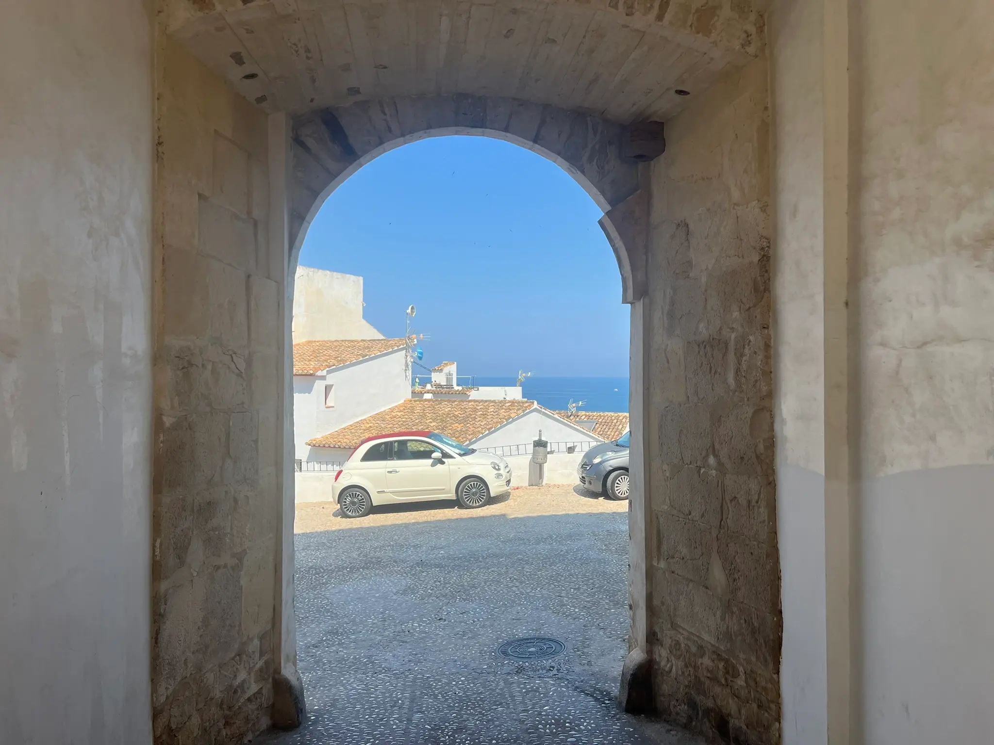 A view through the historic stone archway of Portal Vell in the old town of Altea, Spain. The arch frames a sunlit cobblestone square with white houses, a parked white compact car, and the blue Mediterranean Sea in the distance.