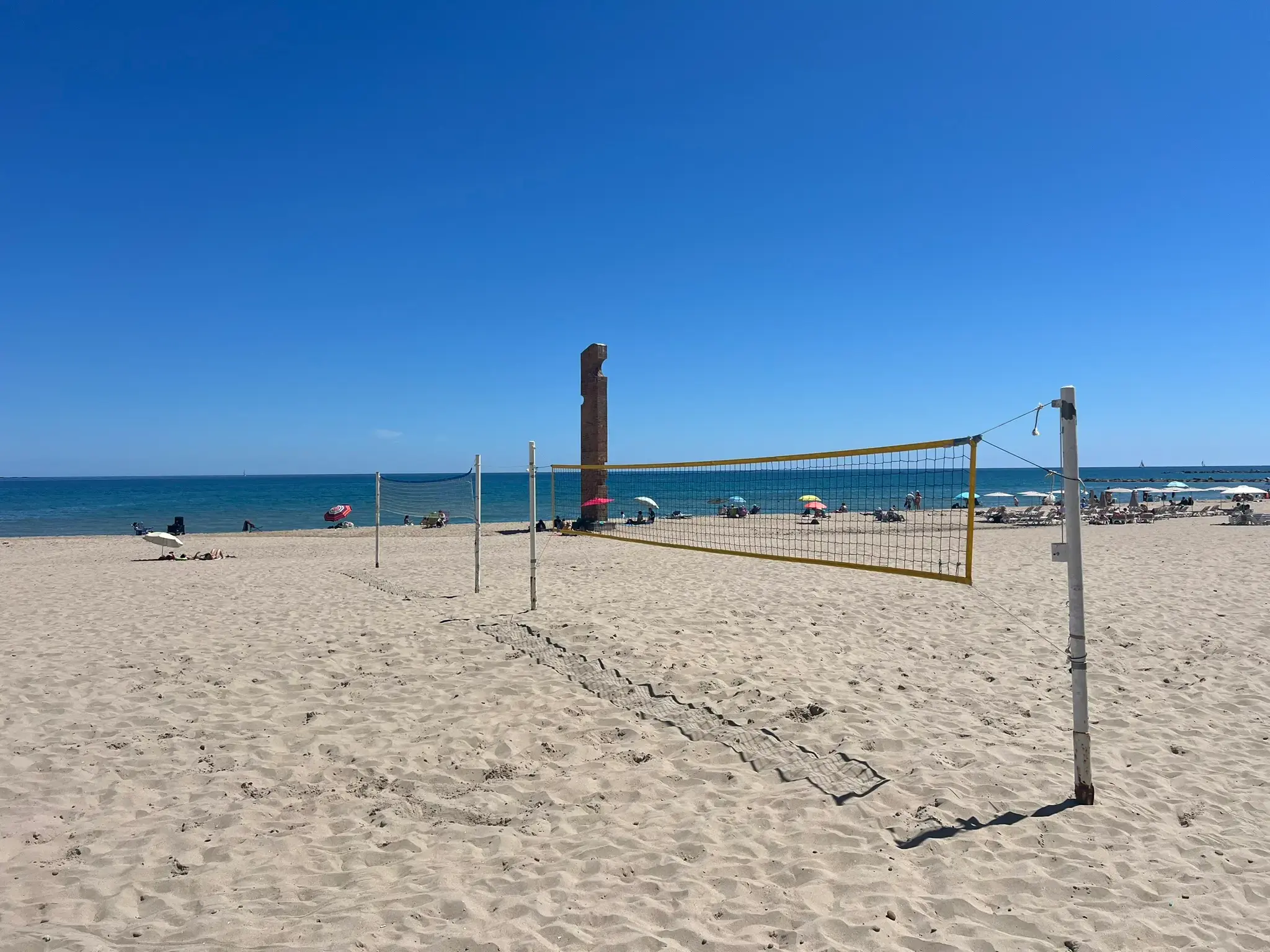 Beach volleyball court on Playa Carrer la Mar in El Campello. A yellow-framed net is set up on the wide sandy beach under a clear blue sky, with the Mediterranean Sea and several beach umbrellas visible in the background.