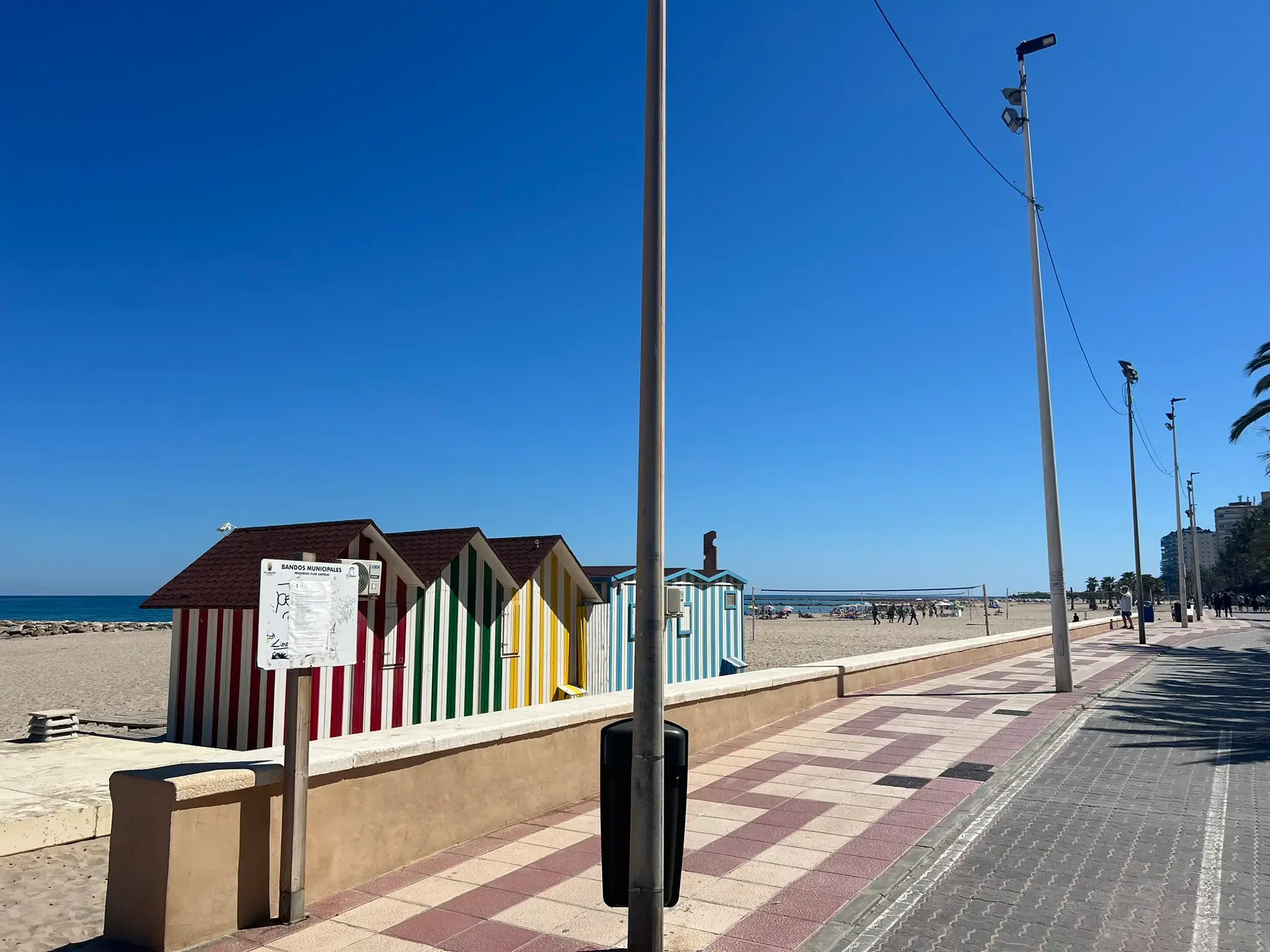 A row of iconic colorful striped beach huts on the sandy shore of El Campello. The small wooden cabins feature red, green, yellow, and blue vertical stripes against a bright blue sky. A paved promenade with a patterned tile walkway and tall street lamps runs alongside the beach area.