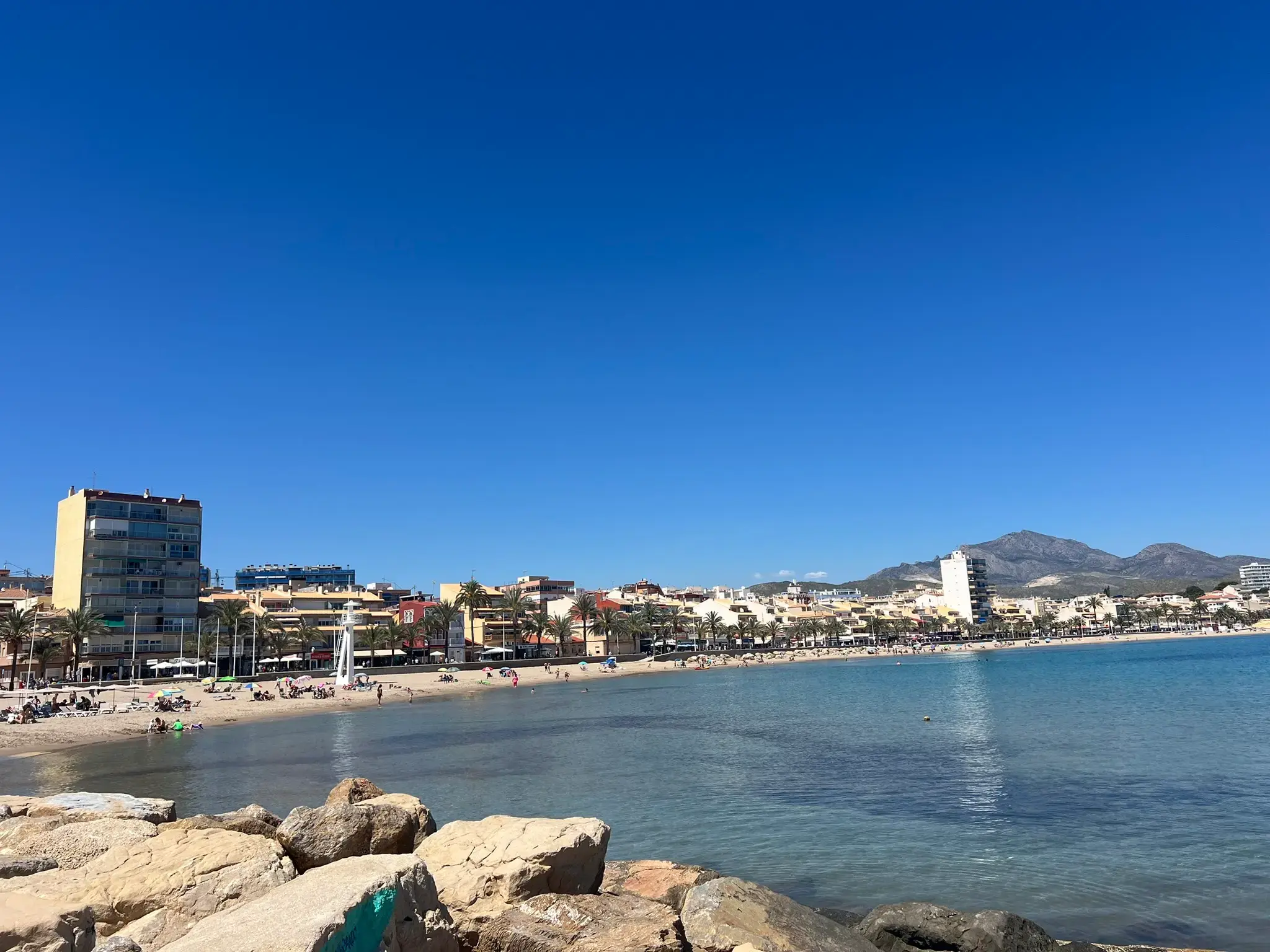 Wide coastal panorama from El Campello, Alicante, showing the turquoise Mediterranean bay, sandy beach, and distant mountain peaks of the Costa Blanca region under a clear sunny sky.