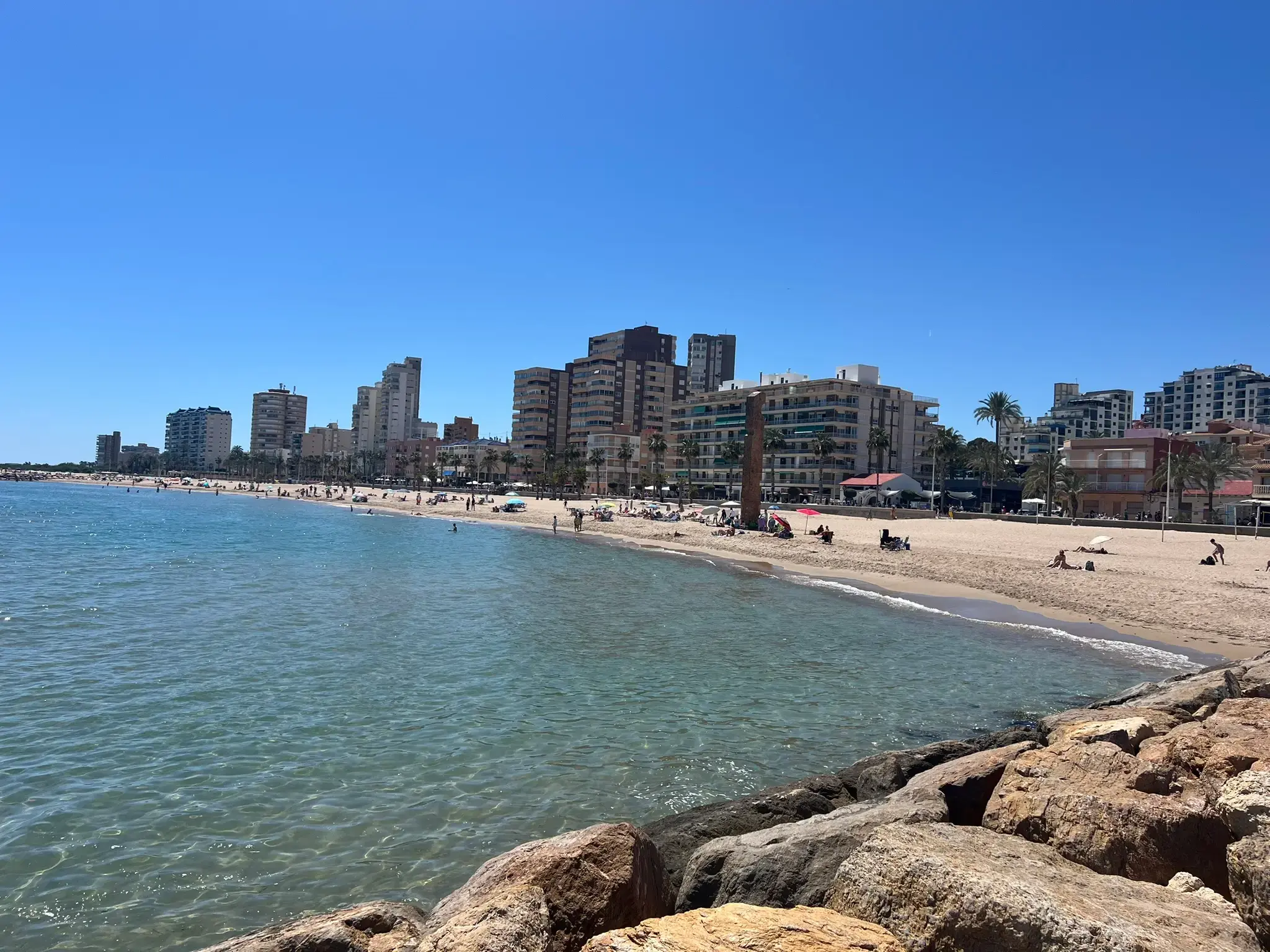 A rock breakwater at Platja del Carrer la Mar in El Campello on a bright, sunny day. The camera looks from the water along the central path of the boulders towards the wide sandy beach. The waterfront is lined with multiple modern high-rise apartment and hotel buildings under a clear blue sky. A single person is walking near the edge of the rocks, and distant beachgoers are visible.