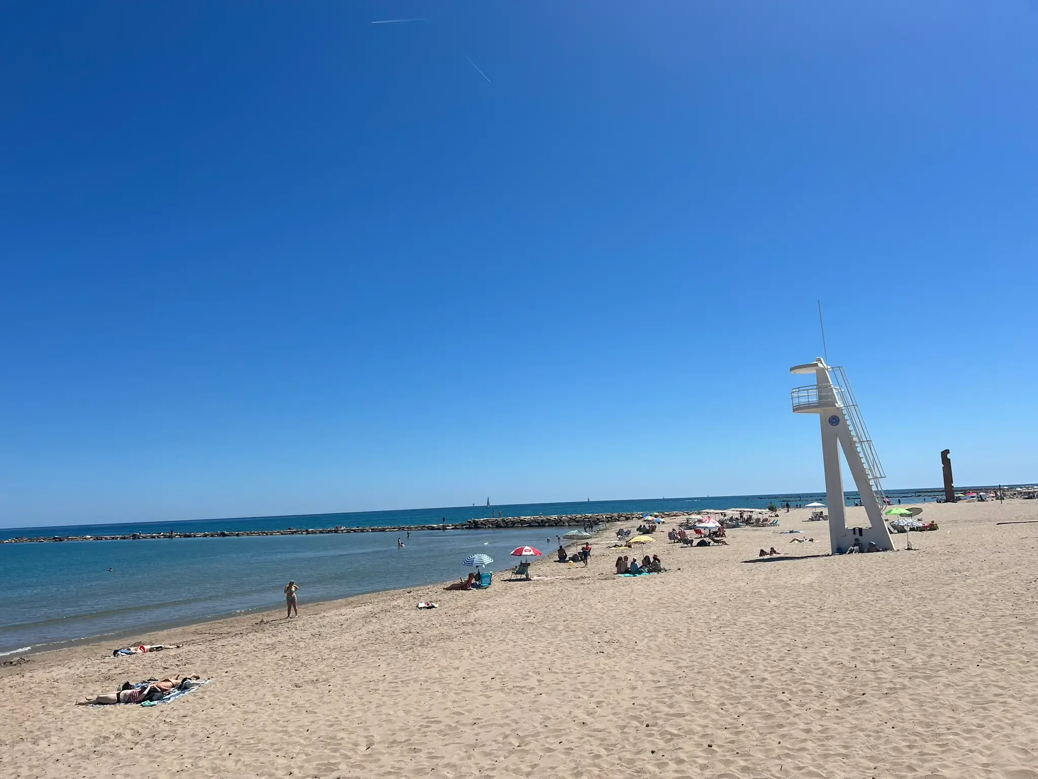 A tall white lifeguard tower on the wide sandy beach of Playa Carrer la Mar in El Campello. Tourists are relaxing under umbrellas near the calm blue Mediterranean Sea, which is protected by a stone breakwater under a cloudless sky.