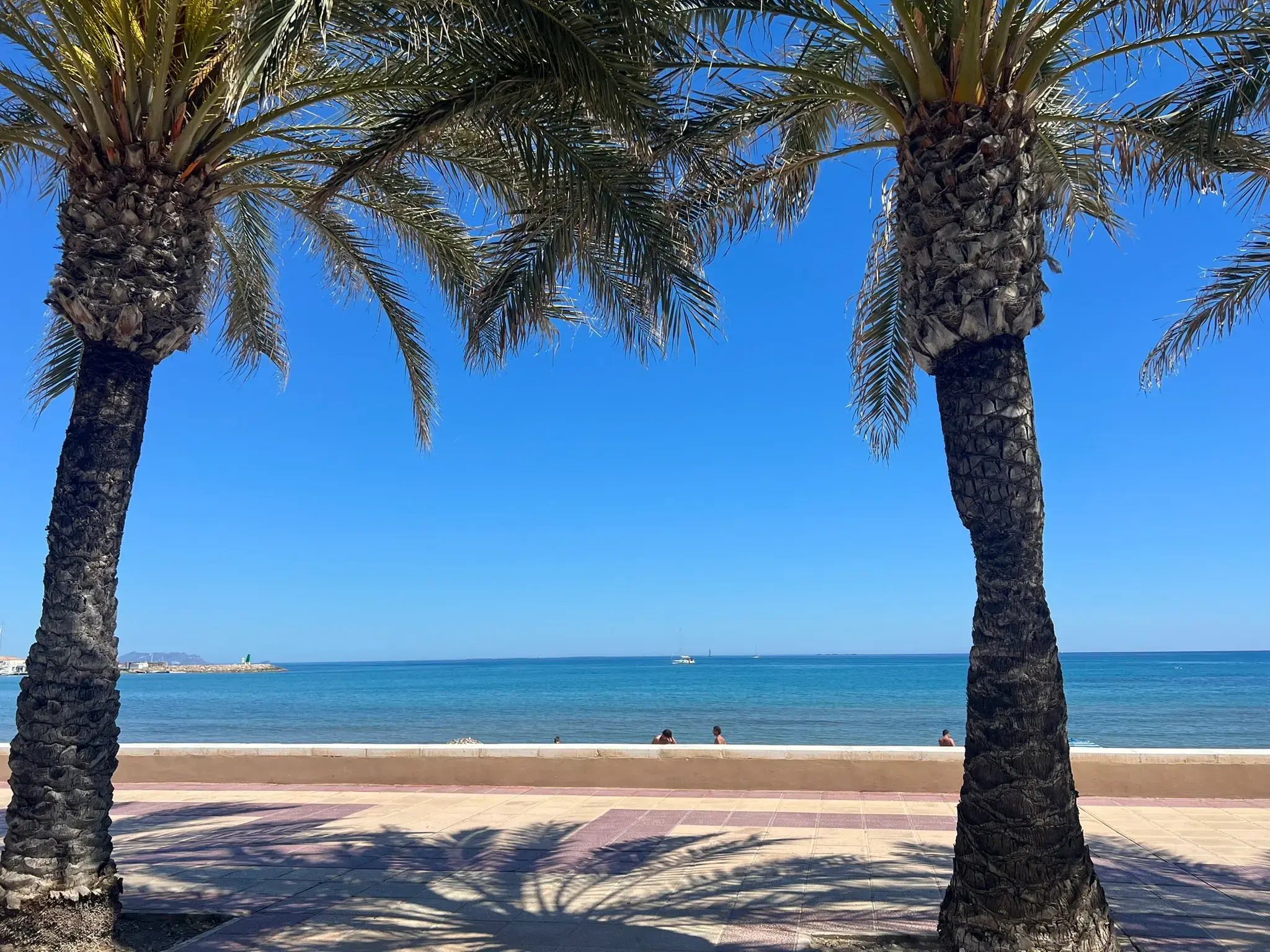 A white modern lifeguard tower stands on the sandy Playa Carrer la Mar in El Campello. People are sunbathing and swimming in the calm Mediterranean Sea, which is protected by a stone breakwater under a vast clear blue sky.