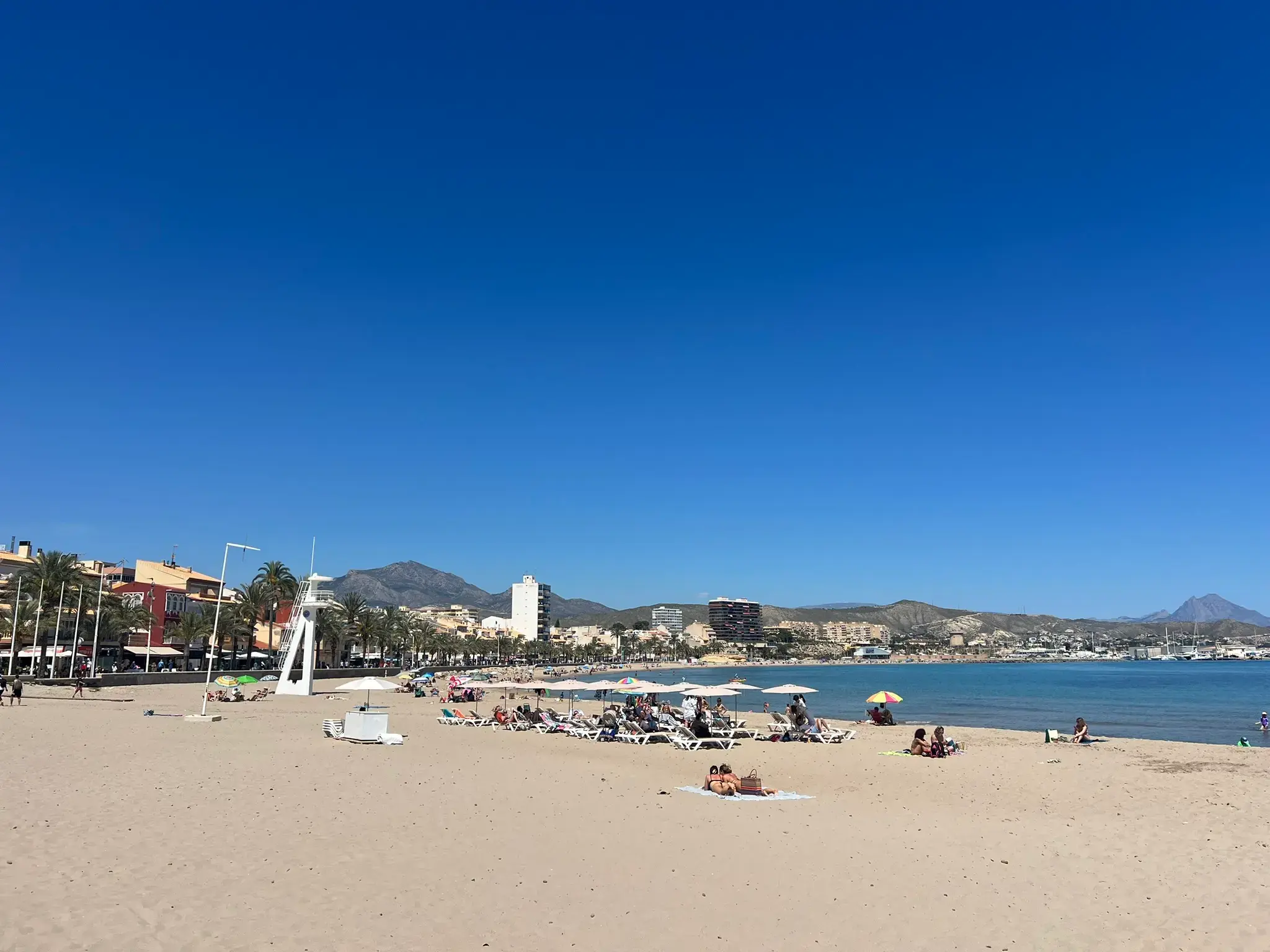A lively afternoon at the sandy beach in El Campello. The photo captures small waves washing onto the shore, scattered beachgoers enjoying the sun, and the modern city skyline featuring a tall brown monument tower against a cloudless blue sky.