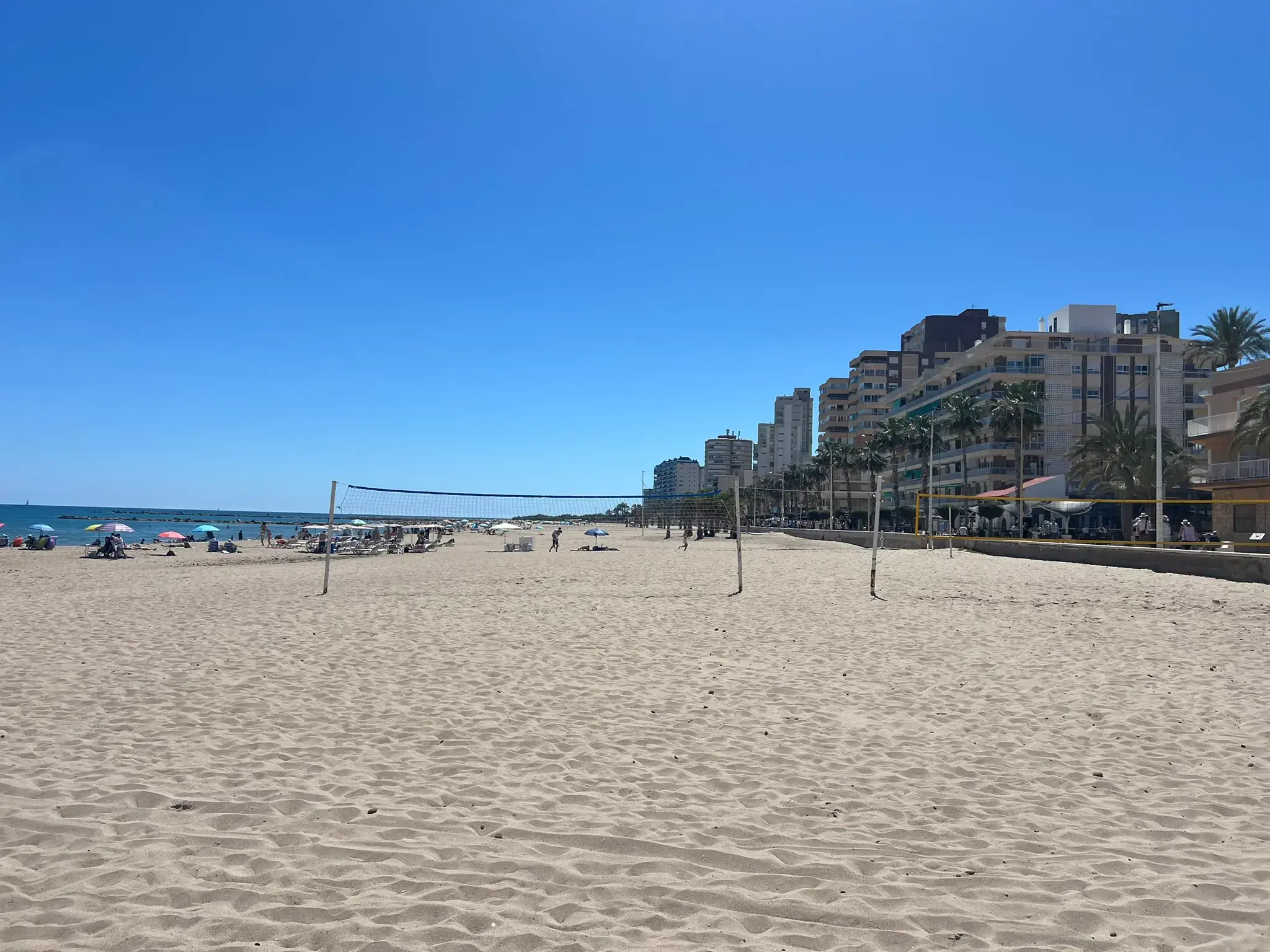 Wide sandy beach area in El Campello featuring volleyball nets. The beach is scattered with colorful umbrellas and sunbathers, bordered by a long row of coastal residential buildings and palm trees under a sunny blue sky.