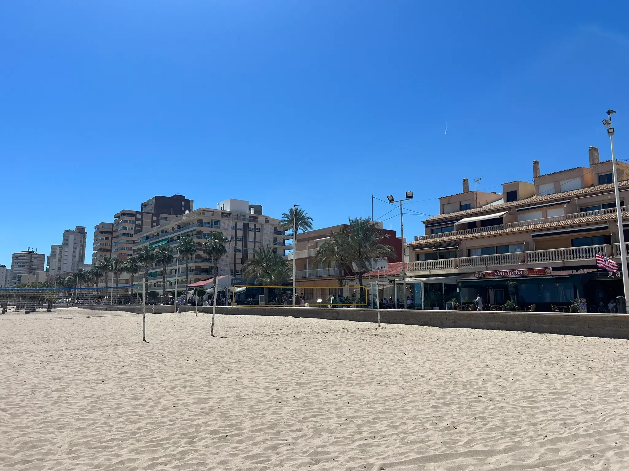 Sunny pedestrian promenade in El Campello featuring palm trees casting long shadows on the patterned tile walkway. The scene shows beachgoers, local shops, and the sandy beach under a bright blue sky.