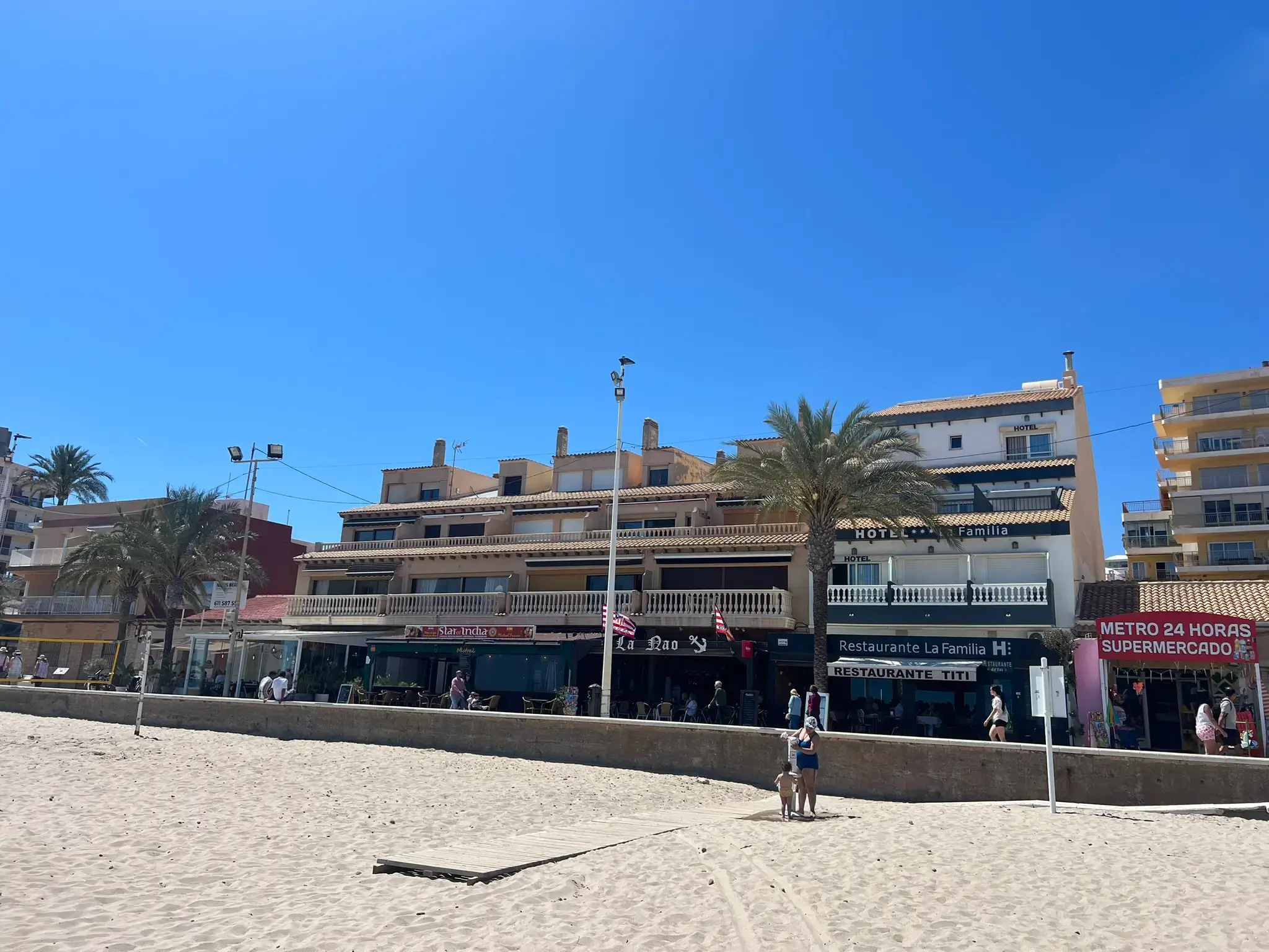 Beachfront view of Carrer la Mar in El Campello. The image shows the sandy beach in the foreground and a row of coastal buildings featuring Restaurante La Familia, Restaurante Titi, and a 24-hour supermarket under a clear blue sky.