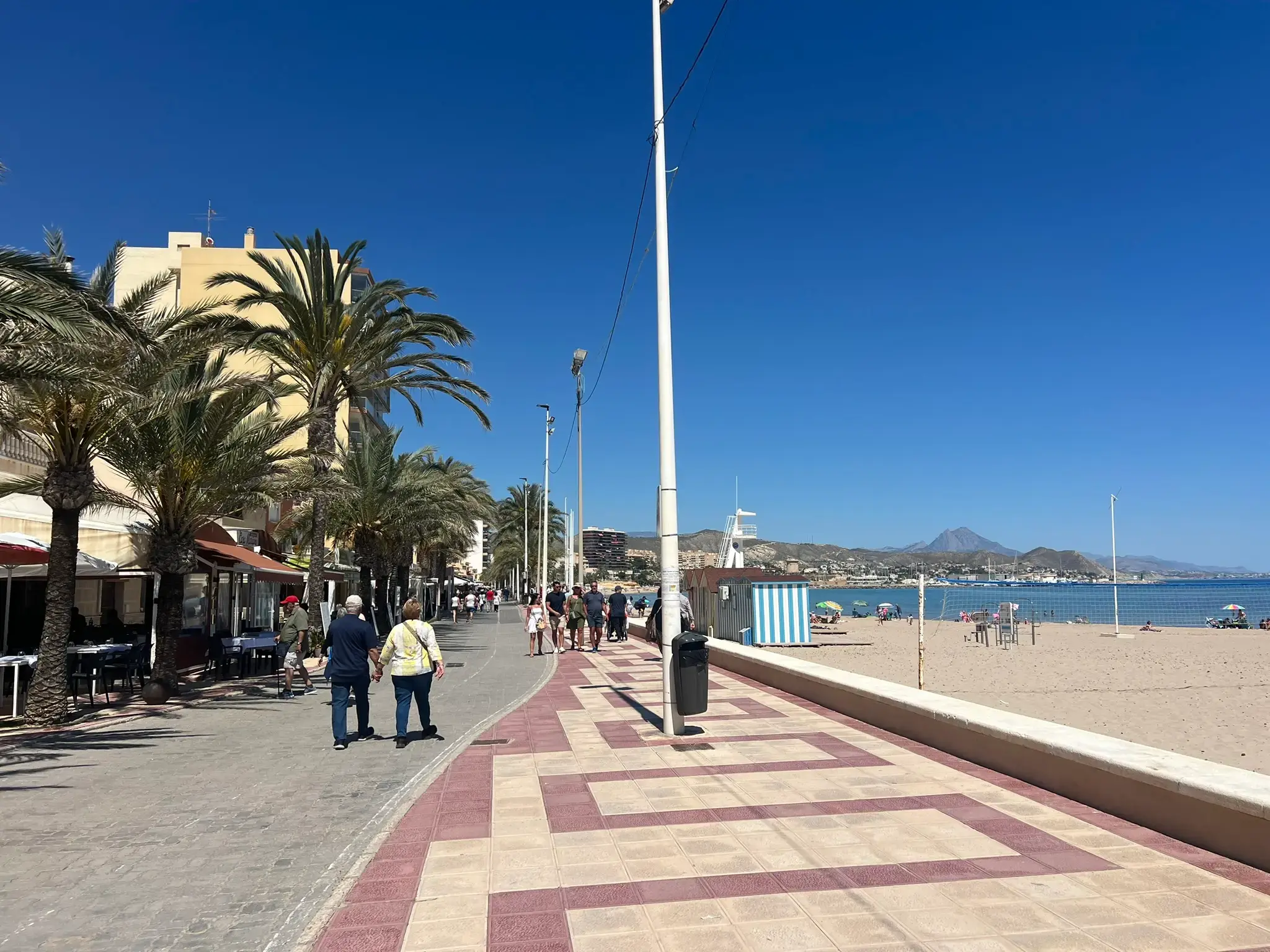 Beachfront promenade in El Campello, Alicante, featuring outdoor cafes, palm trees, and pedestrians. The wide sandy shore of Playa Carrer la Mar and distant mountain peaks are visible under a bright blue sky.