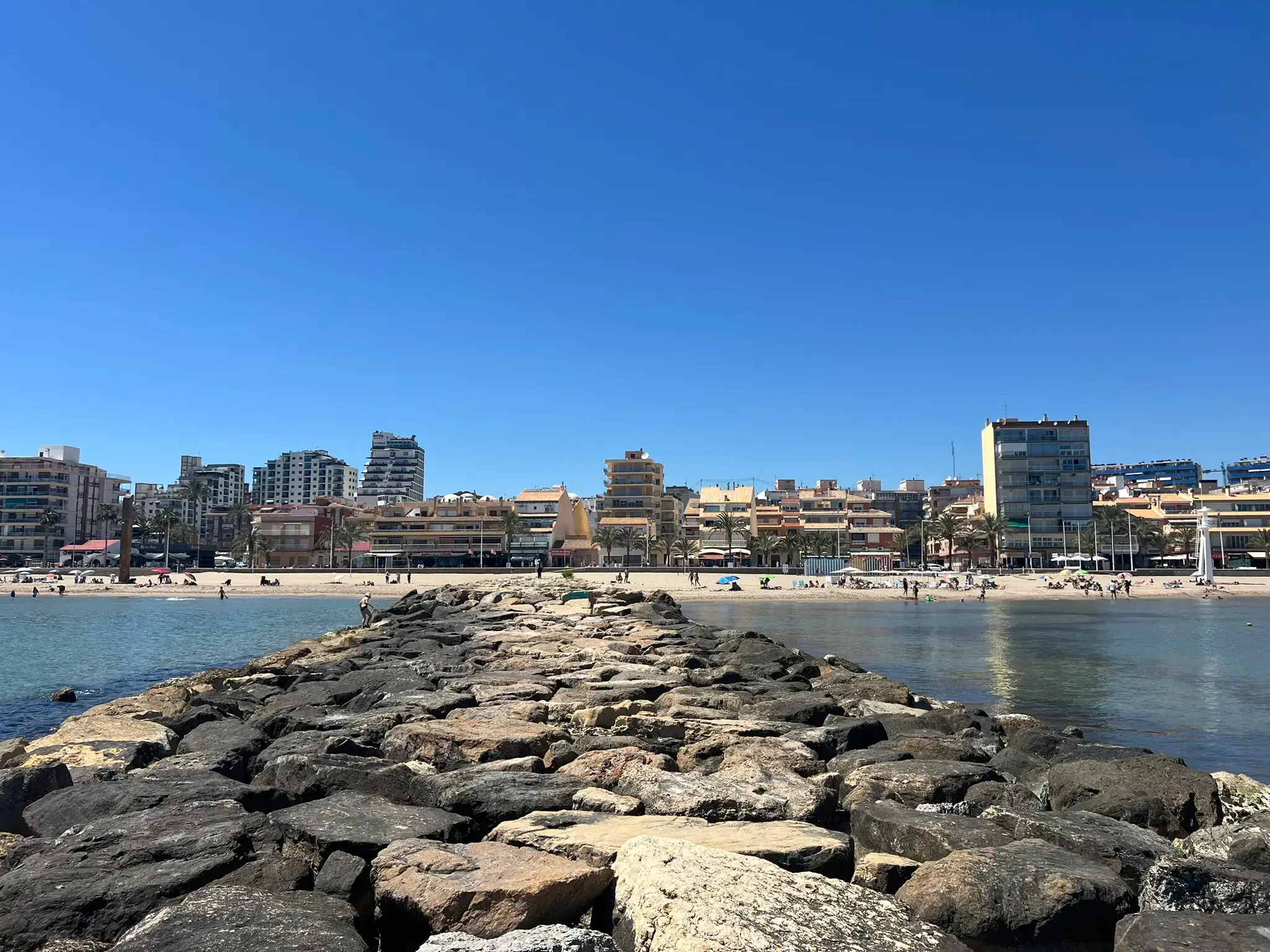 Wide-angle view of the El Campello coastline from a rocky breakwater. The turquoise Mediterranean Sea flows towards a sandy beach populated with sunbathers. The background features a long line of modern high-rise apartments and hotels under a clear, deep blue sky.
